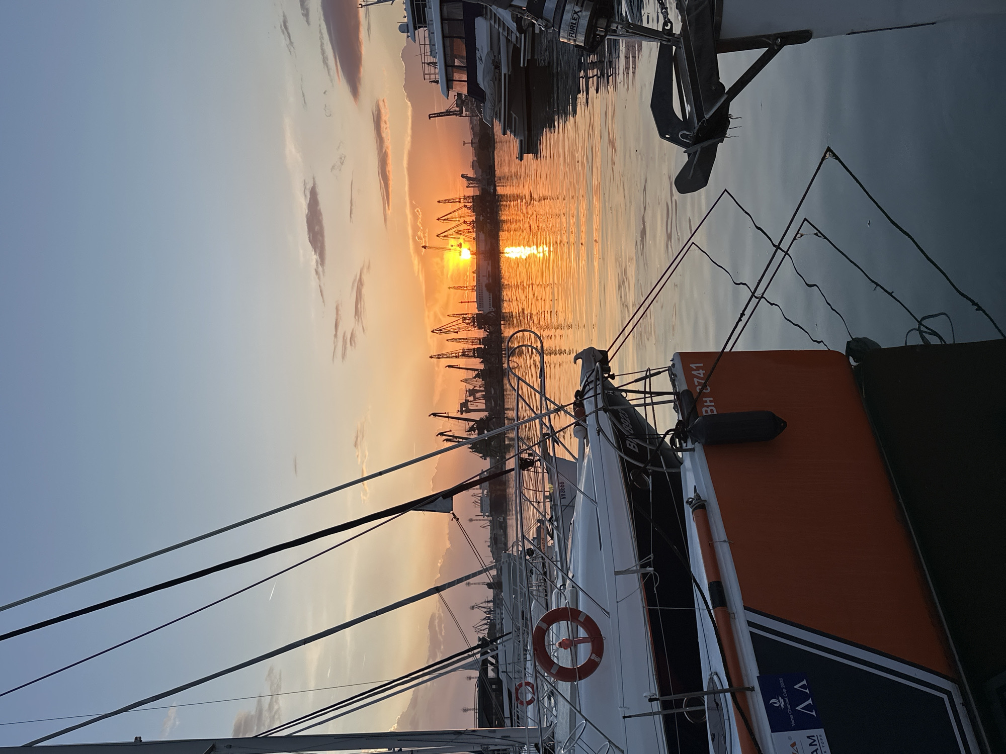 The image shows a sunset over the Varna port in Bulgaria, viewed from a boat. In the background, the sun is setting behind a line of harbor cranes, reflecting intensely on the water. Several boats are docked in the foreground, including one with an orange hull section and a life preserver. Rigging lines stretch from the foreground boat towards the sky. The sky is a mix of blue and orange hues due to the sunset.