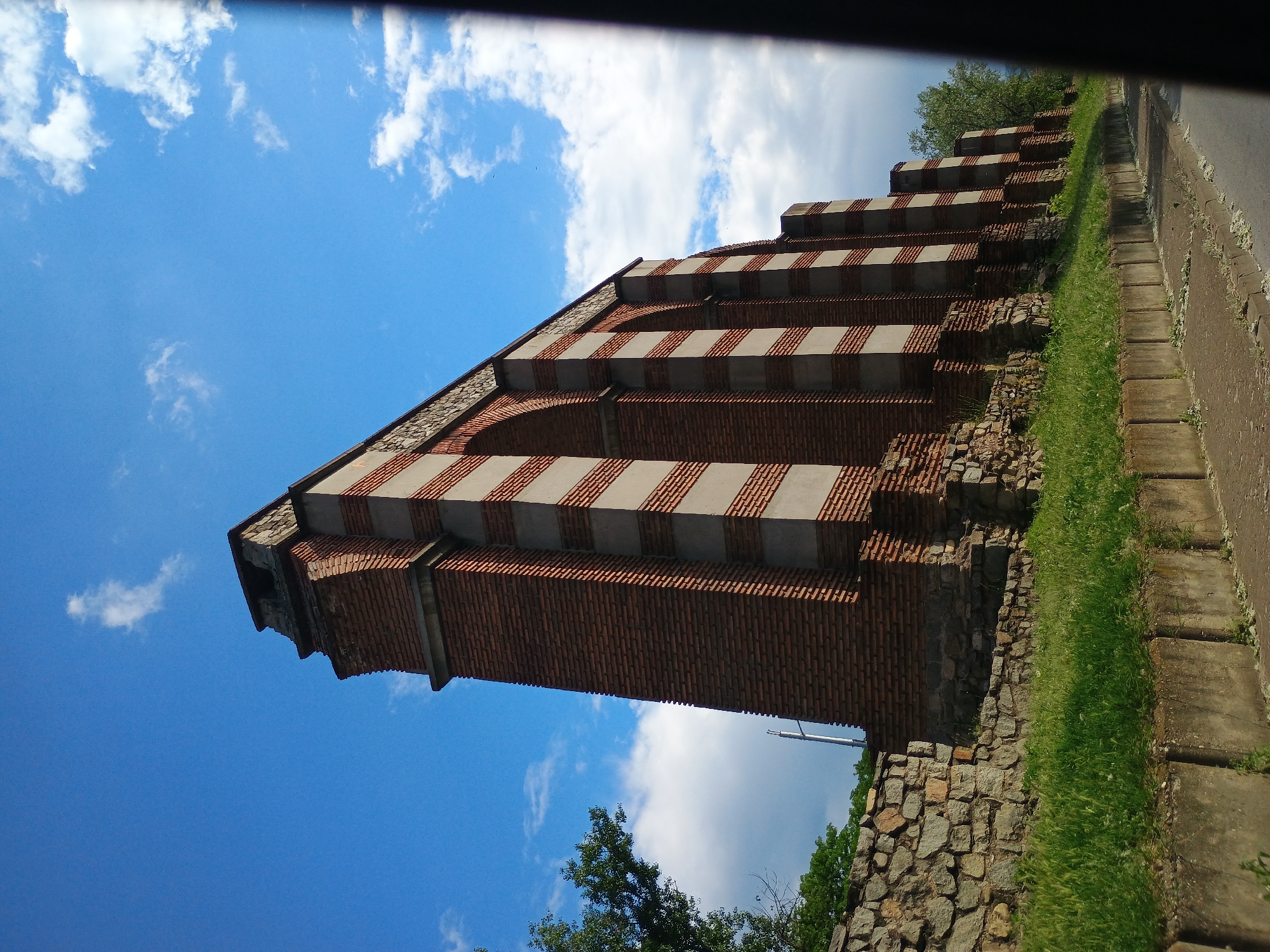 The image captures a tilted view of a brick structure with alternating red brick and gray bands, set against a backdrop of blue sky with scattered clouds. The structure appears to be part of a larger ruin, with a stone base and remnants of walls or foundations nearby. The ground level is obscured by vegetation, including grass and some trees. A concrete curb or sidewalk runs along the right edge of the image. The image's perspective suggests it was taken looking upwards from a low angle. The location is an unknown city, in an unknown region.