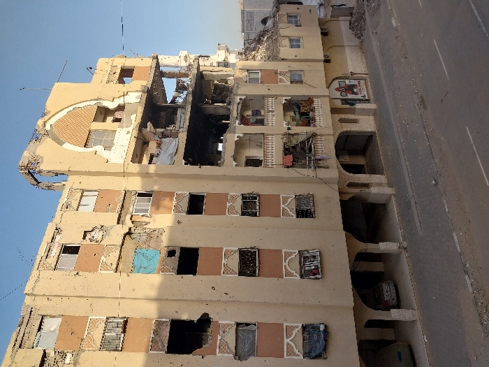 The image displays a multi-story residential building located in Khan Yunis, Palestinian Territory, exhibiting extensive damage. The building, constructed with a light beige facade accented by horizontal orange bands, shows numerous impact marks consistent with bullet holes and shrapnel distributed across its entire front.
Significant structural collapse is evident on the upper floors of the right side, exposing interior rooms, displaced concrete, and twisted rebar. A prominent decorative arched feature at the top right of the building is severely compromised. Many windows are shattered or entirely missing, while some are boarded with materials, including a blue tarp on the third floor from the bottom on the left. Damaged metal security grates are present on several window openings. Balconies on the right side are partially destroyed, with clothing items visible hanging from an intact section of one balcony. The roofline is uneven, showing debris and partial collapse.
The ground floor features an open area beneath arched pillars, functioning as a parking space where a silver sedan and another covered vehicle are present. A single commercial sign with a red logo is visible on the far right of the ground level. The foreground includes an asphalt road with white lane markings, flanked by a paved sidewalk covered with scattered debris and dirt. The sky above is clear and blue. Another partially visible, seemingly damaged structure is present in the background to the far right. No individuals are visible in the scene.