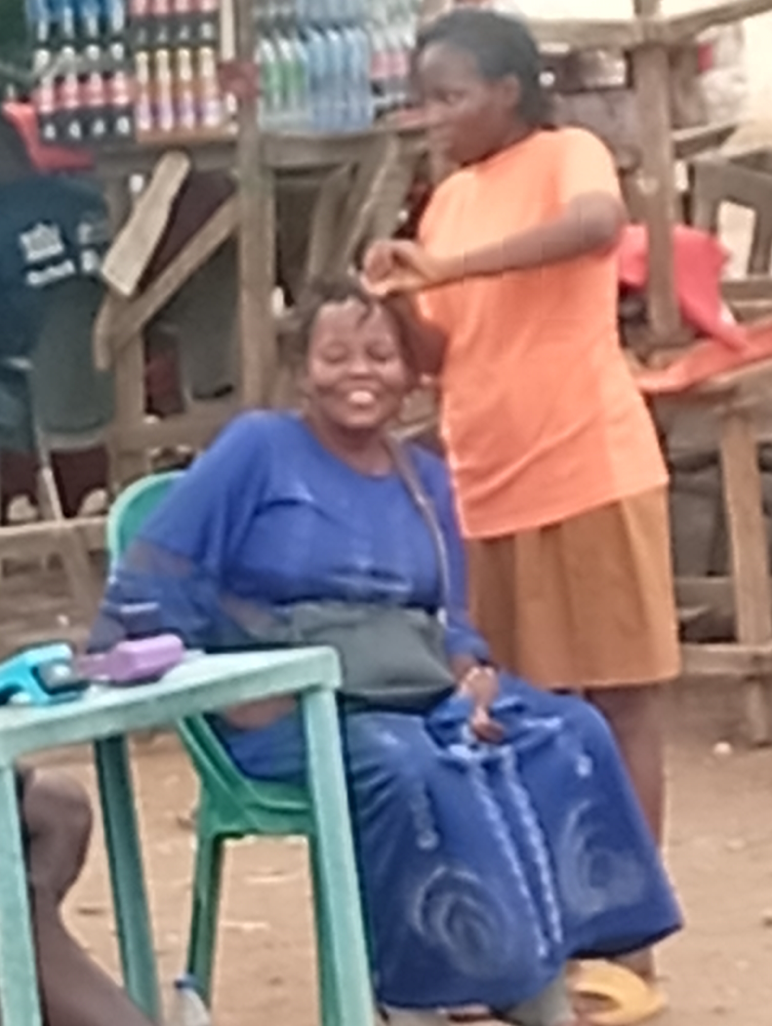 In Sabon Tasha, Nigeria, an outdoor scene captures two women interacting. One woman, seated in a green plastic chair, wears a blue patterned dress, has a green purse, and smiles as the other woman styles her hair. The standing woman is dressed in an orange shirt and a tan skirt. A light green table sits in front of the seated woman, holding objects including a phone and clippers. In the background, a variety of bottled and canned goods are visible on wooden shelving units. The setting appears to be an informal, possibly roadside, hair styling location.