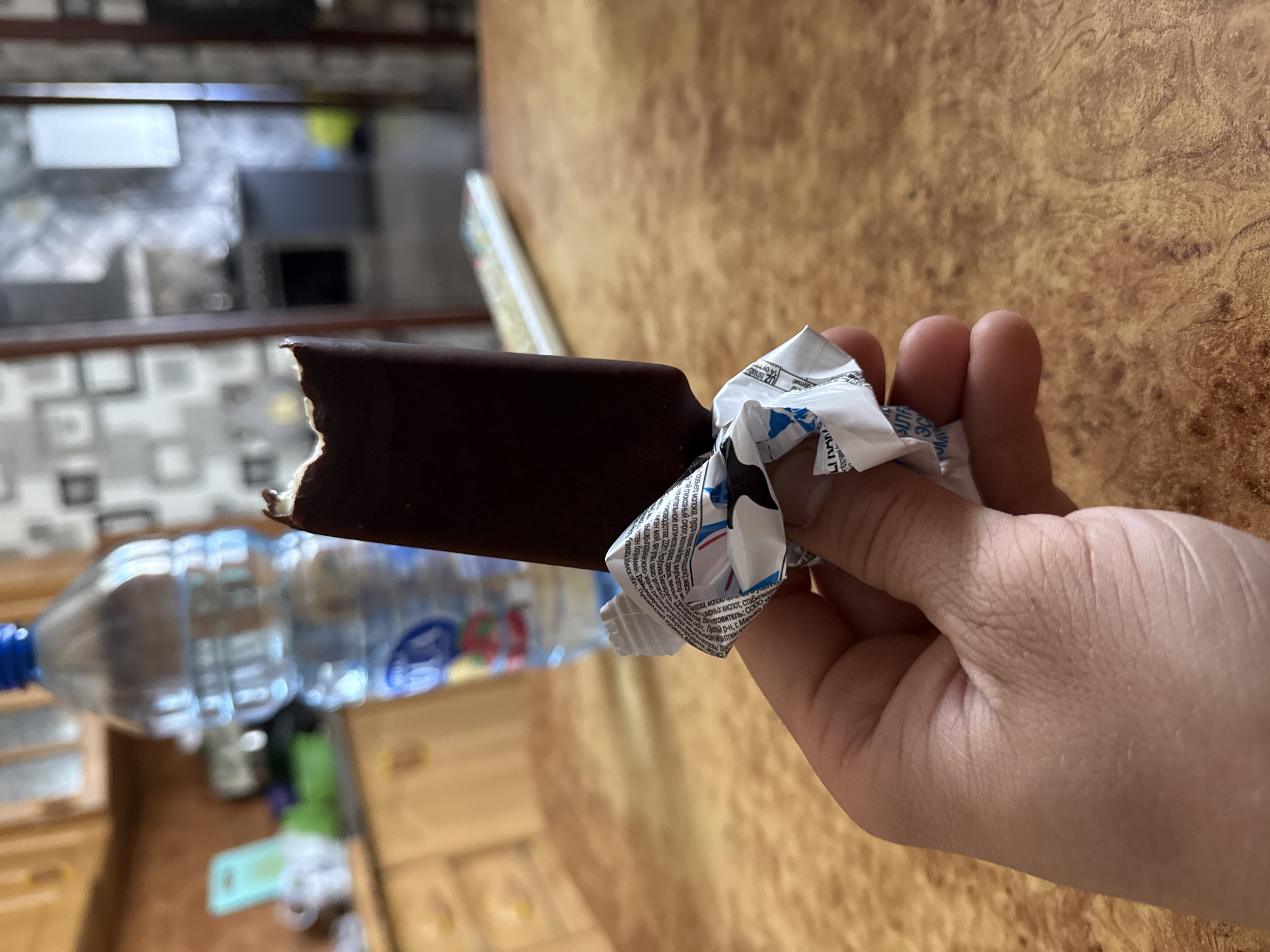 A hand holds a partially eaten chocolate-covered ice cream bar, still in its wrapper. A water bottle is visible in the background. The scene is likely indoors on a surface with a wood-like pattern. OCR suggests the ice cream is manufactured in Maryina Horka, Belarus.