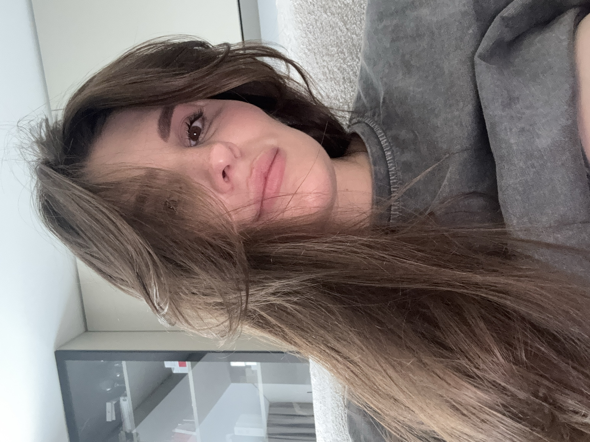 This close-up selfie depicts a young woman, presented in an image rotated 90 degrees clockwise, suggesting she was lying down when the photo was taken. She has long, wavy dark brown hair that cascades around her face and extends across the lower part of the frame. Her dark eyes are open, and her expression is soft and relaxed, with her lips slightly parted. She has well-defined eyebrows and a fair complexion.
The woman is wearing a casual, long-sleeved grey shirt or sweatshirt. The fabric has a faded or stone-washed appearance, with visible stitching along the collar or shoulder area, indicating a comfortable and well-worn garment.
The background suggests an indoor setting. To her right (if viewing the image as taken upright), a light grey, textured fabric object, likely a sofa or cushion, is visible. To her left, there's a light-colored wall and a dark, reflective rectangular object, possibly a screen, mirror, or framed picture, showing indistinct reflections of an indoor space. There are no clear location cues within the image itself to confirm the provided context of Sofiivska Borshchahivka, Ukraine. The lighting is soft, and no specific time of day or weather conditions can be determined. No text is visible in the media file.
