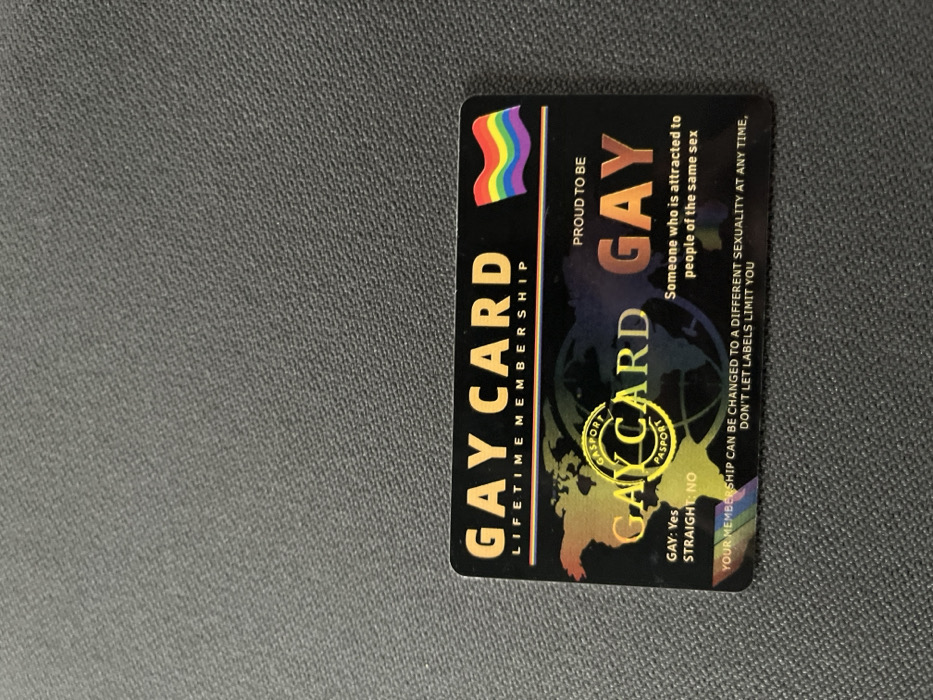 A black, rectangular novelty card, labeled "GAY CARD LIFETIME MEMBERSHIP," is resting on a dark grey, textured fabric surface in Maslarevo, Bulgaria. The card features a rainbow flag emblem in the upper right corner. Below the main title, a stylized holographic world map is present, overlaid by a circular yellow seal marked "GASPORT PASPORT." Textual elements on the card include "GAY: Yes" and "STRAIGHT: NO" on the left side. On the right, "PROUD TO BE GAY" is displayed, followed by the definition "Someone who is attracted to people of the same sex." The bottom of the card contains a statement: "YOUR MEMBERSHIP CAN BE CHANGED TO A DIFFERENT SEXUALITY AT ANY TIME, DON'T LET LABELS LIMIT YOU," presented over a subtle rainbow gradient. No individuals or actions are depicted in the scene.