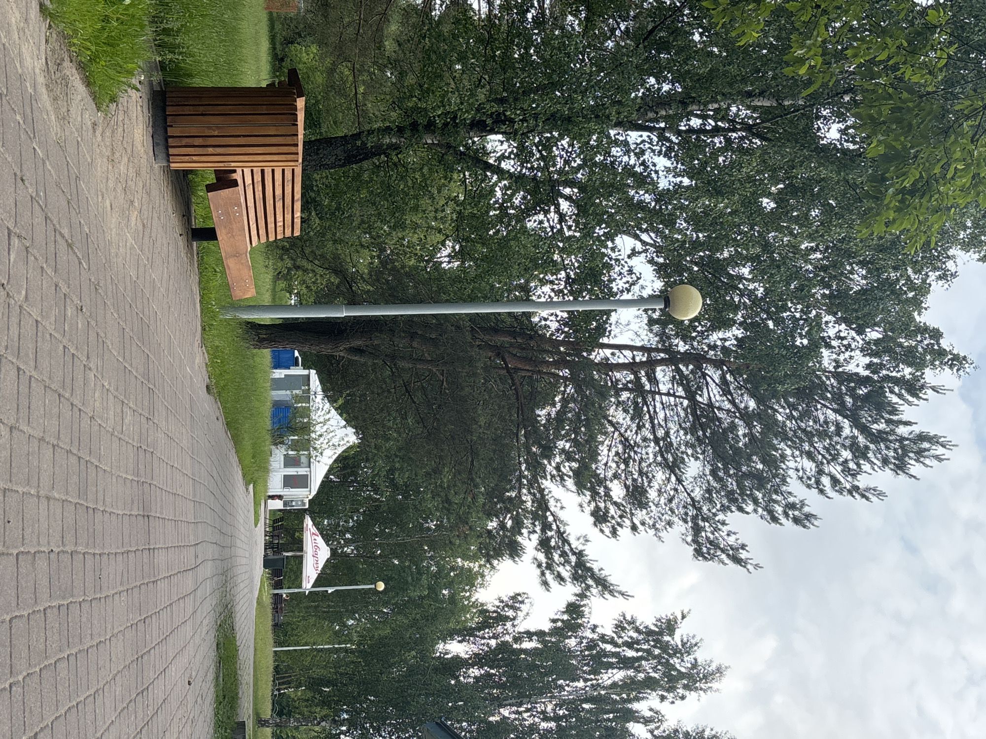 The image shows a paved walkway in a park-like setting. On the left side of the walkway is a modern wooden bench with a trash receptacle integrated into its design. A tall lamppost with a spherical light fixture stands further down the walkway. In the background, there are several tents, one of which appears to be a white structure with glass panels, possibly a cafe or pavilion. Another tent has a white canopy with red lettering that appears to read "Любарка". Tall trees, including birches and pines, surround the area, suggesting a natural or semi-natural environment. The sky is overcast. The overall impression is a public outdoor space, possibly a recreational area or a park. Based on the visible signage and the context of public parks in Minsk, Belarus, this scene is likely located within Minsk.