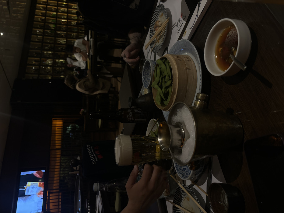 A dimly lit table displays Japanese food and drinks. Edamame in a wooden steamer basket sits beside a small patterned bowl. Tempura remnants fill a white plate near a Suntory menu. A white bowl holds a sauce with a spoon. A metal sake server filled with ice is beside a beer glass. A hand pours liquid from a clear bottle. The scene suggests a relaxed dinner in a restaurant setting.