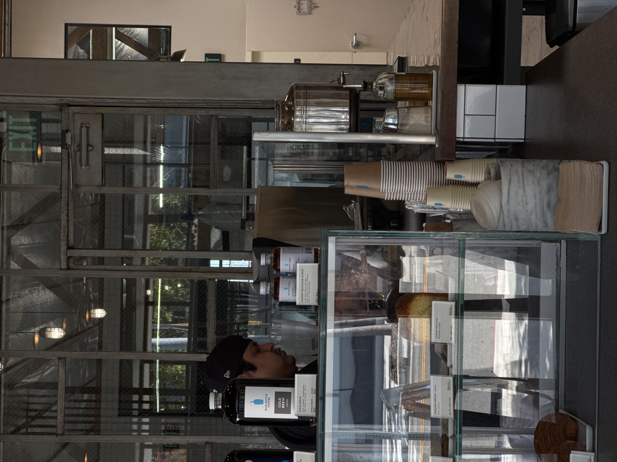Here is a description of the image:
The image captures the interior of a Blue Bottle Coffee shop. A man wearing a black baseball cap stands near a glass display case filled with baked goods such as lemon matcha pound cake and banana bread. He is positioned in front of shelves containing bottles of Blue Bottle Cold Brew Coffee and Cherry Blossom Syrup. Behind him, a window with a metal frame offers a glimpse of the outside. An exit sign is visible above the window frame. Coffee cups are stacked on a shelf above the glass display case.