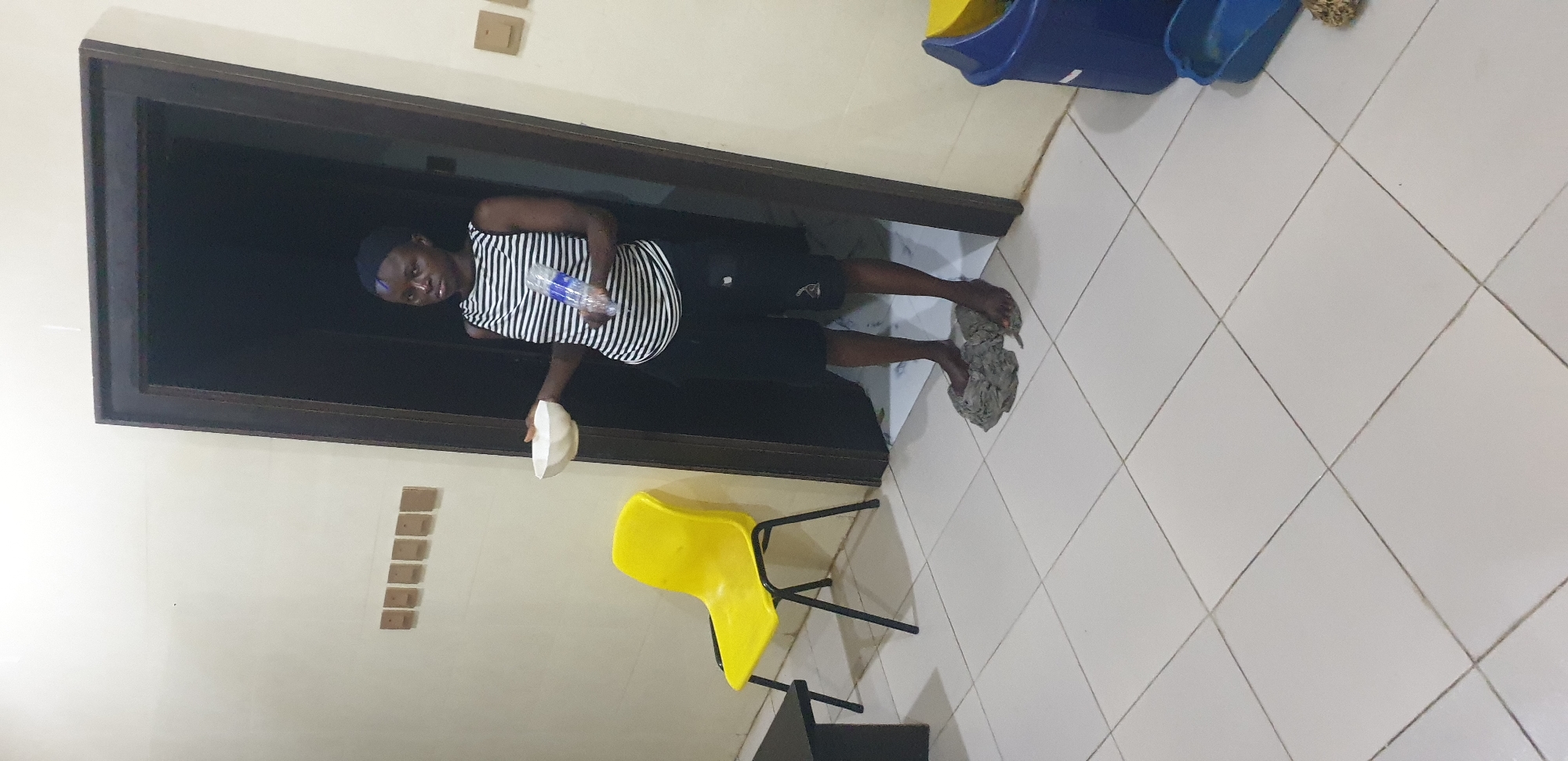 This image depicts an indoor scene in the city of Barden Yana, Nigeria. An adult individual, appearing female, with dark skin and short, dark hair, stands centered in a dark wooden doorway. She is wearing a sleeveless top with black and white horizontal stripes and dark shorts. Her bare feet are positioned on two grey, crumpled fabric items on the light-colored tiled floor, which has grey grout lines arranged in a grid pattern. The individual holds a white bowl in her left hand and a clear plastic water bottle with a blue label in her right hand. To the left of the doorway, a yellow plastic chair with black metal legs is visible, along with a partial view of a dark rectangular table in the foreground. Six rectangular, light brown wall plates are mounted horizontally on the wall to the left of the doorway. Two similar light brown wall plates are mounted on the wall to the right of the doorway. A blue and yellow plastic bin is partially visible on the far right, with a smaller blue plastic tray on the floor beside it. The area beyond the doorway is dark, indicating a dimly lit space.