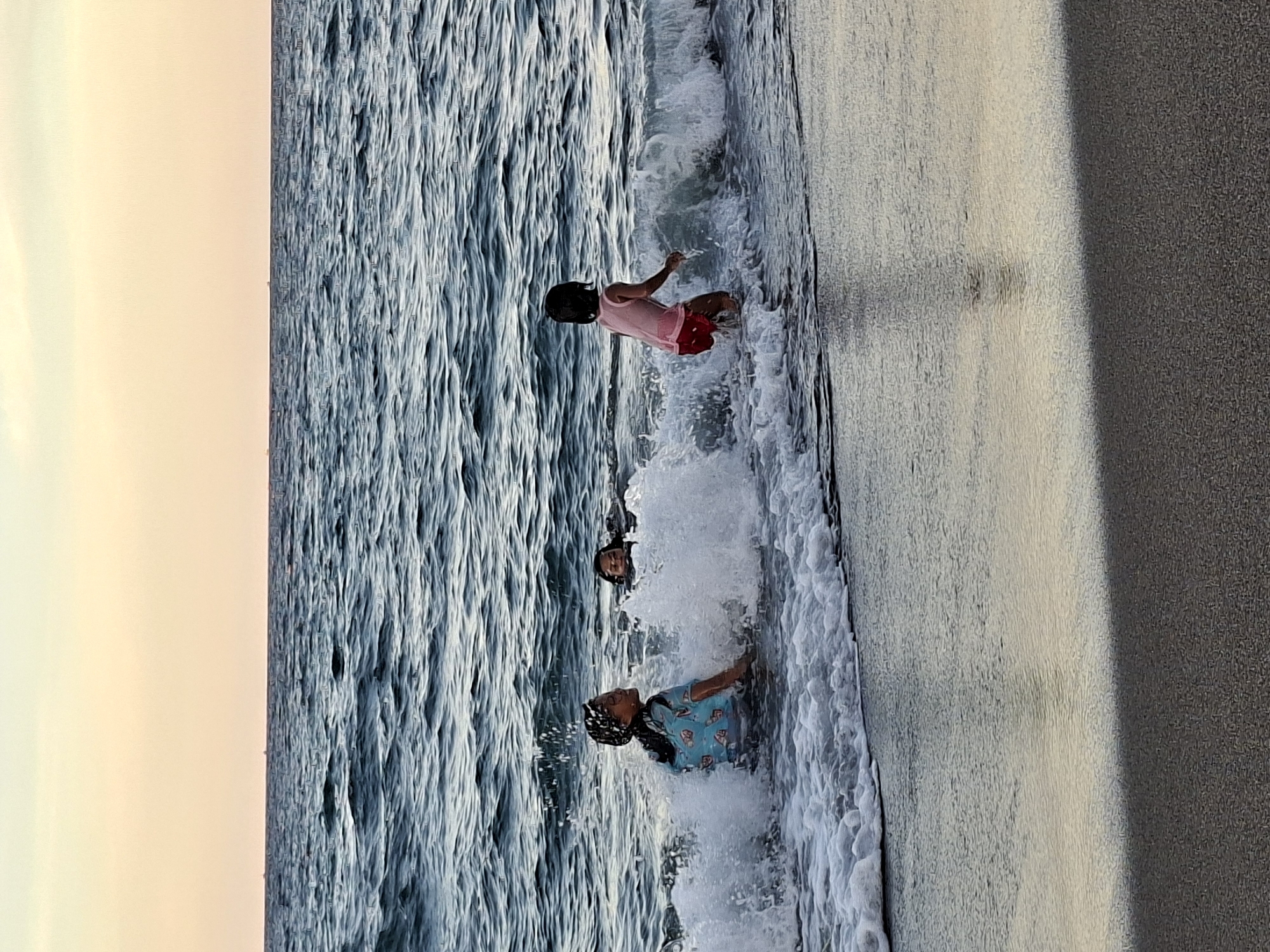Three children are playing in the surf on a beach. The location is Lhokseumawe, Indonesia. One child wears a pink top and red shorts, standing in the water. Another child is submerged in the water up to their shoulders. The third child wears a blue patterned shirt and is wading in the water. Waves are breaking on the shore. The sky is a pale orange. Sand is visible on the beach to the right, with a darker area in the foreground.