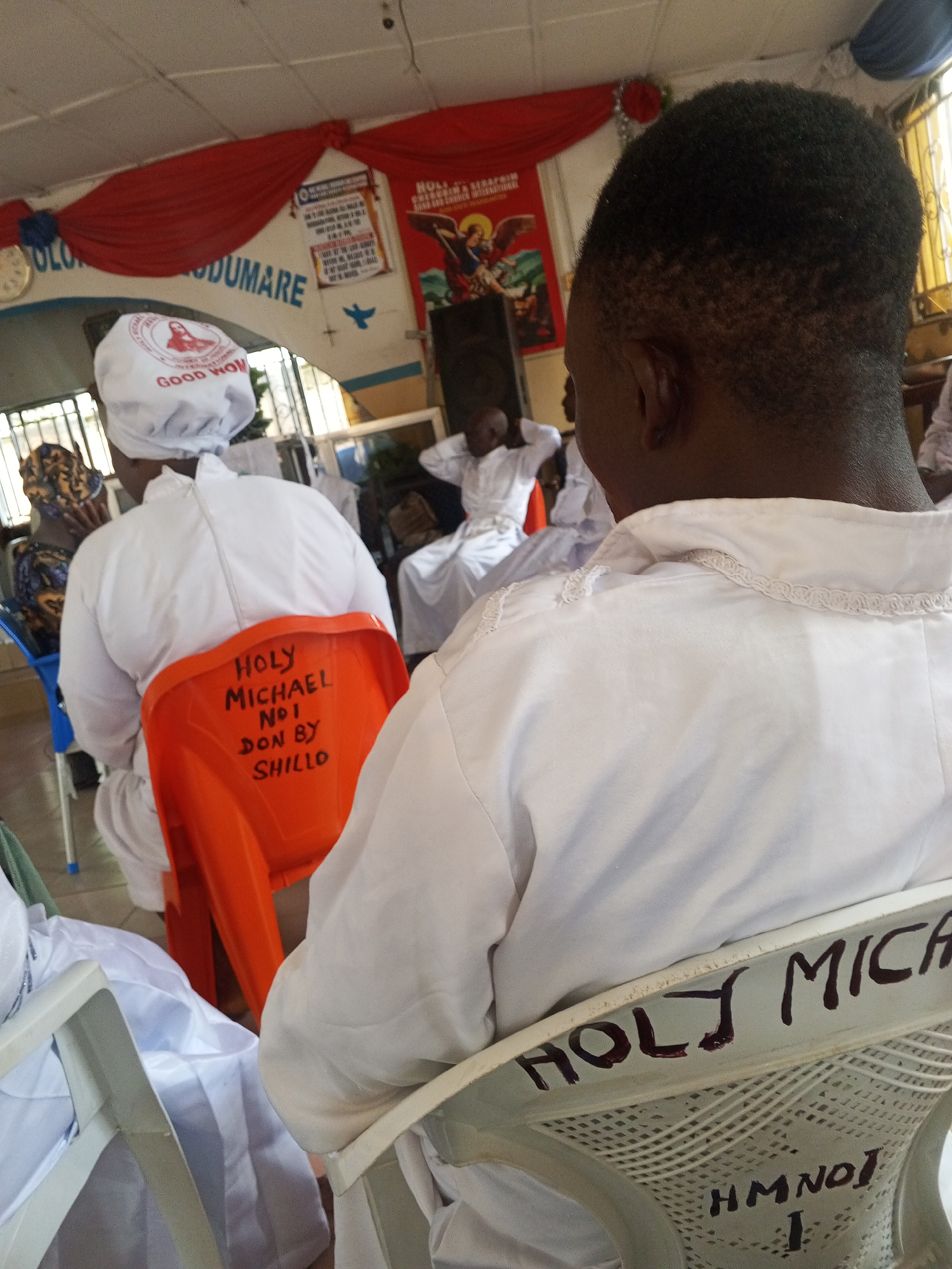 This image depicts an indoor gathering, reportedly in Lokoja, Nigeria, characterized by religious iconography and uniform attire. The perspective is from behind the head of an individual with short, dark, cropped hair, who is seated in a white plastic chair. The back of this chair features handwritten text: "HOLY MICHA" and "HM NO1 I."
In the mid-ground, another individual, also dark-skinned, is seen from behind, wearing a white long-sleeved garment and a white head covering. The head covering has a circular red emblem with an image and the text "GOOD WOMAN." This person is seated in a prominent orange plastic chair, the back of which is inscribed with black marker text: "Holy Michael No1 Don By Shillo."
Multiple other individuals are partially visible in the background, mostly seated and wearing white or patterned head coverings. Windows with vertical security bars are visible in the background, admitting natural light.
The walls are decorated with red fabric drapes across the top, and a curved light-blue banner displaying white text, partially legible as "OLOLO GODUMARE," along with a blue bird silhouette. A white clock with a dark rim is mounted on the wall. To the right, various framed posters and notices are visible, including a large poster depicting a winged figure (likely Archangel Michael) engaged in combat with a dragon, with text such as "SERAPHIM" and "HOLY MICHAEL" discernible. The ceiling is a white drop-tile design. The floor appears to be light-colored.