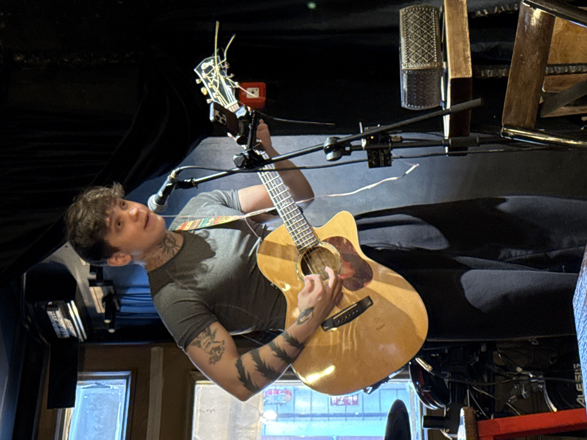 A man with short, dark hair and tattoos on his arms and neck is playing an acoustic guitar on a stage. He is wearing a gray t-shirt and is positioned in front of a microphone and a guitar stand that holds a mobile phone. The background includes dark curtains, lighting equipment, and glimpses of what appears to be the exterior of a building. Based on the text seen through the window, specifically the word "Түбәтәй", the location is Kazan, Russia.