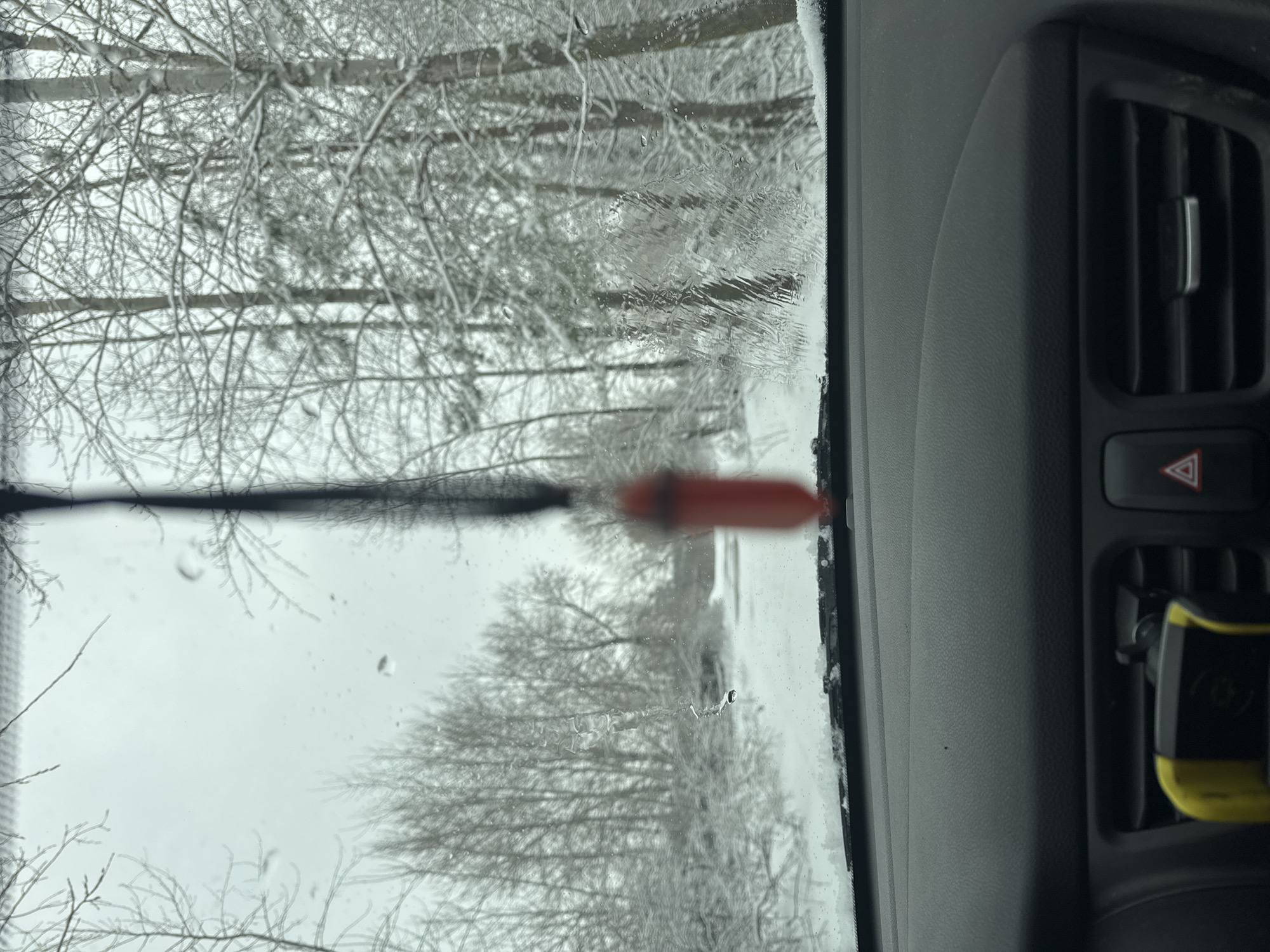 The image appears to be taken from inside a vehicle, looking out through the windshield. The windshield is partially covered with ice or snow. Outside the vehicle, trees with bare branches are visible, suggesting a winter setting. The scene is likely in a snowy environment, as indicated by the general white and gray tones. The presence of ice on the windshield indicates it is likely cold weather, possibly below freezing temperatures. Inside the vehicle, a dashboard element is visible on the right side of the frame, including a hazard light button. A red-handled tool is seen partially visible in front of the windshield.
Without further information about the location within the city of Chaikovsky, Russia, a precise location cannot be determined, but the landscape suggests a wooded or park area, typical of many urban environments in Russia.