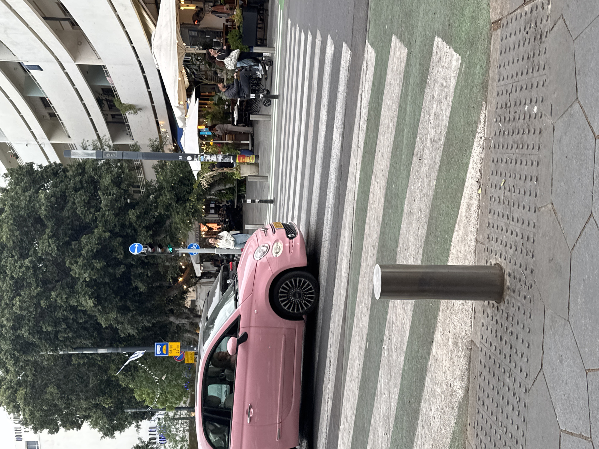 A pink Fiat 500 car is parked on a crosswalk in Tel Aviv, Israel. The crosswalk has a green bicycle lane adjacent to it. The vehicle partially obstructs the pedestrian crossing. Nearby are trees and buildings, including restaurants with outdoor seating. A metal bollard is installed near the crosswalk on the sidewalk. A traffic light pole and signs are also visible. Pedestrians are present in the background.