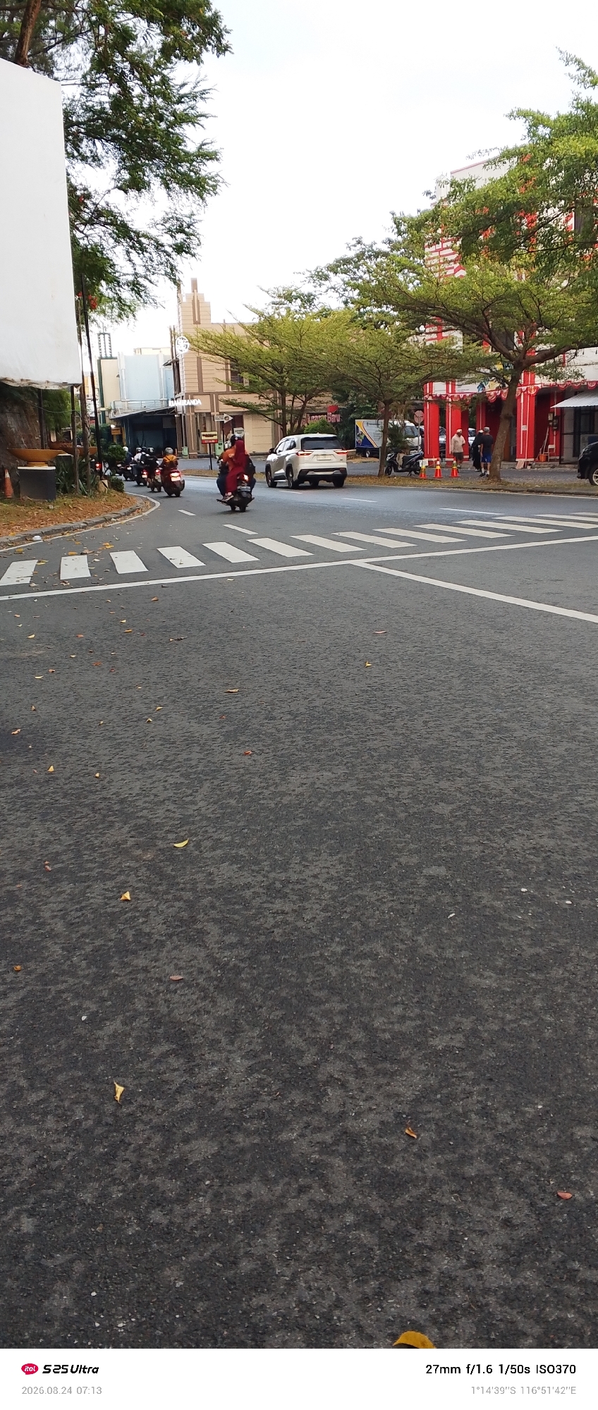 The image captures a street scene in Balikpapan, Indonesia. In the foreground, the asphalt road displays scattered dry leaves and white pedestrian crossing markings. The midground shows a white SUV driving away from the camera, and several motorcycles are moving along the road. One motorcycle in particular has two individuals, a driver and a passenger wearing red. To the left, a large white structure is partially visible, with trees and buildings in the background. The buildings are modern in style with some featuring red and white architectural elements. The sky above is bright and overcast. The timestamp on the image indicates it was taken on August 24, 2026, at 07:13. The coordinates pinpoint the location at 1°14'39"S 116°51'42"E.