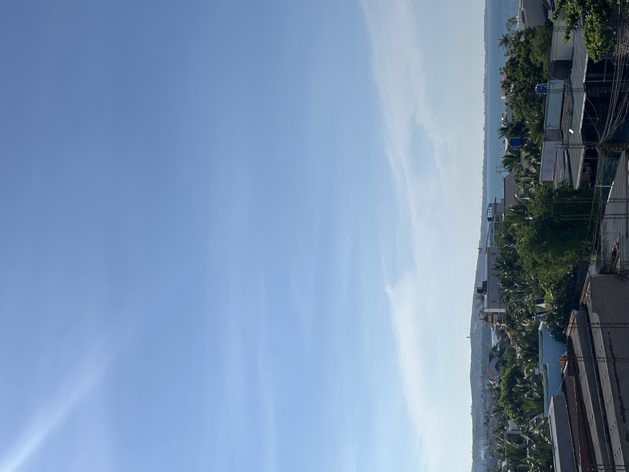 This image captures a panoramic view of the coastal city of Mũi Né, Vietnam. The upper portion of the image is dominated by a clear, bright blue sky with a faint sunbeam streaking from the upper left. Below, the horizon is marked by a distant, hazy coastline with undulating hills, topped by what appears to be a lighthouse or monument.
The middle ground reveals a densely built urban landscape interspersed with lush green tropical vegetation, primarily palm trees. Various buildings, including houses and low-rise structures with flat roofs, are visible. Some buildings have solar panels on their roofs, suggesting modern infrastructure. The architecture appears to be a mix of traditional and contemporary styles, with pastel-colored accents on some rooftops.
The foreground is occupied by the rooftops of buildings under construction or in a state of disrepair. These structures feature exposed concrete, dark roofing materials, and visible construction elements like rebar. Electrical wires are also prominently displayed, indicating active infrastructure.
The overall scene depicts a sunny day in a coastal town, characterized by a juxtaposition of natural beauty and ongoing urban development. The image is a static, wide-angle shot, implying a stationary viewpoint, likely from a higher elevation. No people or animals are discernible in the image. The environment suggests a warm, possibly humid climate.