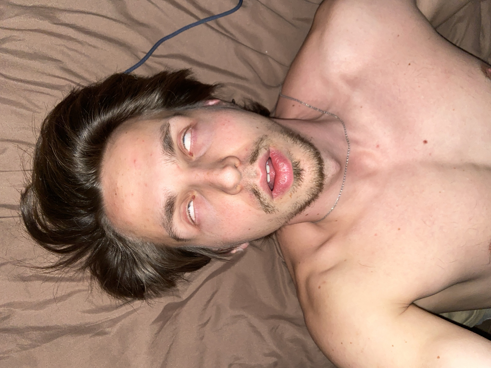 A Caucasian male with brown hair and a light beard is lying on a brown bedsheet. His eyes are rolled back, showing only the whites. His mouth is slightly open. He is shirtless and wearing a silver necklace. A blue cord is visible in the upper left corner of the image. The location in Šušanj, Montenegro is not determinable from the image.