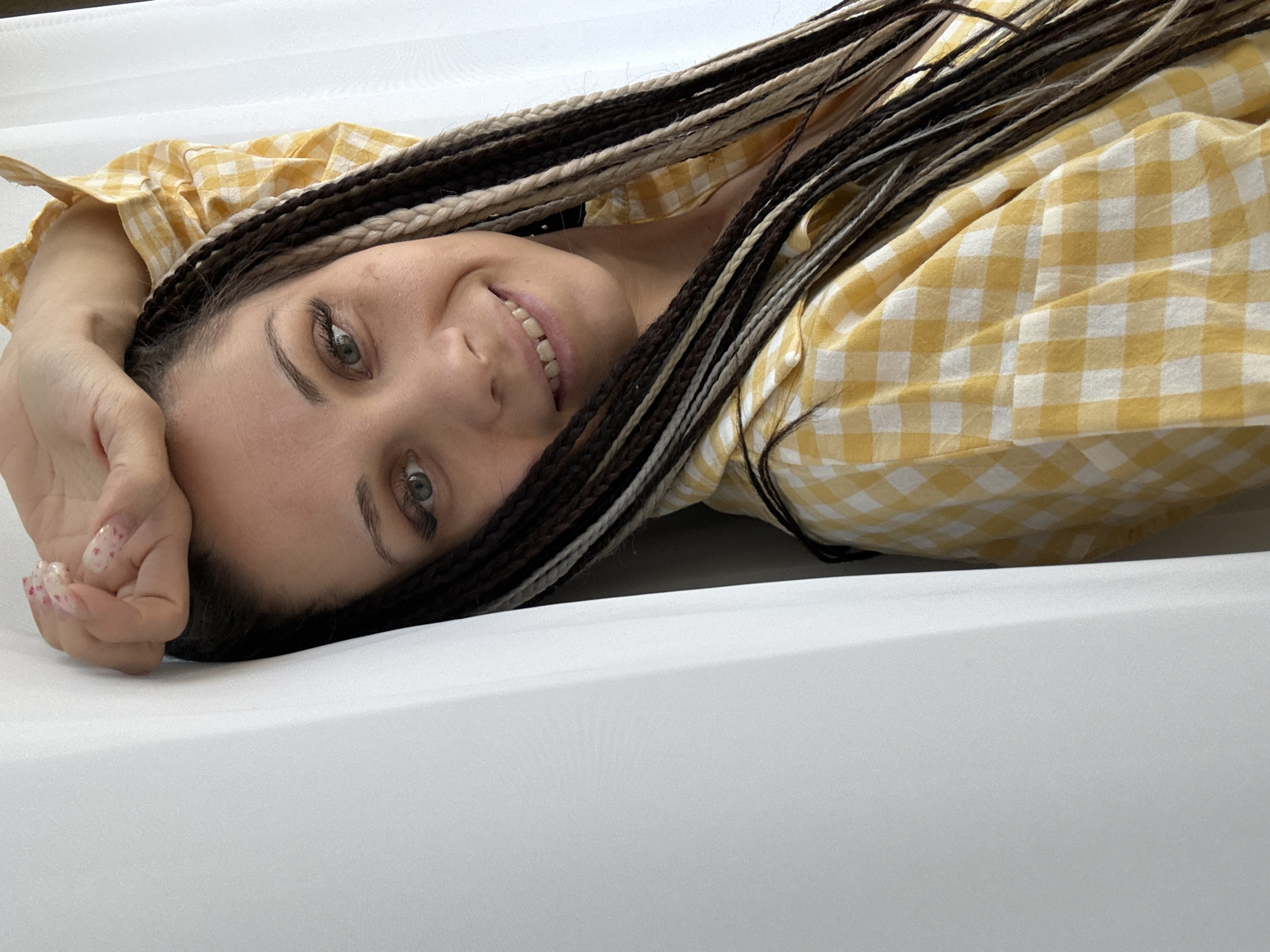 A woman with braided hair and a yellow plaid shirt is lying on a white surface. Her head is tilted, and she is smiling at the camera. Her left hand is near her head. The background is blurry and lacks sufficient detail to establish the location as Rusanovo, Russia.