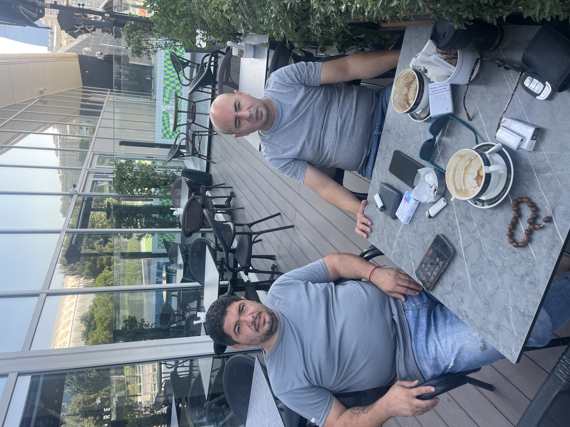 Two men are seated at a table on an outdoor deck, likely at a cafe in Baku, Azerbaijan. The man on the left has dark, curly hair and a beard, and is wearing a gray t-shirt and blue jeans. He has a tattoo on his left arm and is wearing a red bracelet on his right wrist. The man on the right is bald and is wearing a gray t-shirt and blue jeans.
On the marble-topped table are two cups of coffee, sunglasses, a mobile phone, lighters, a pack of cigarettes, a small black wallet, car keys, and a string of brown beads. In the background, through large windows, the cityscape of Baku is visible, including modern buildings and lush greenery. The cafe appears to be situated in a contemporary architectural setting, with a glass facade and outdoor seating area.