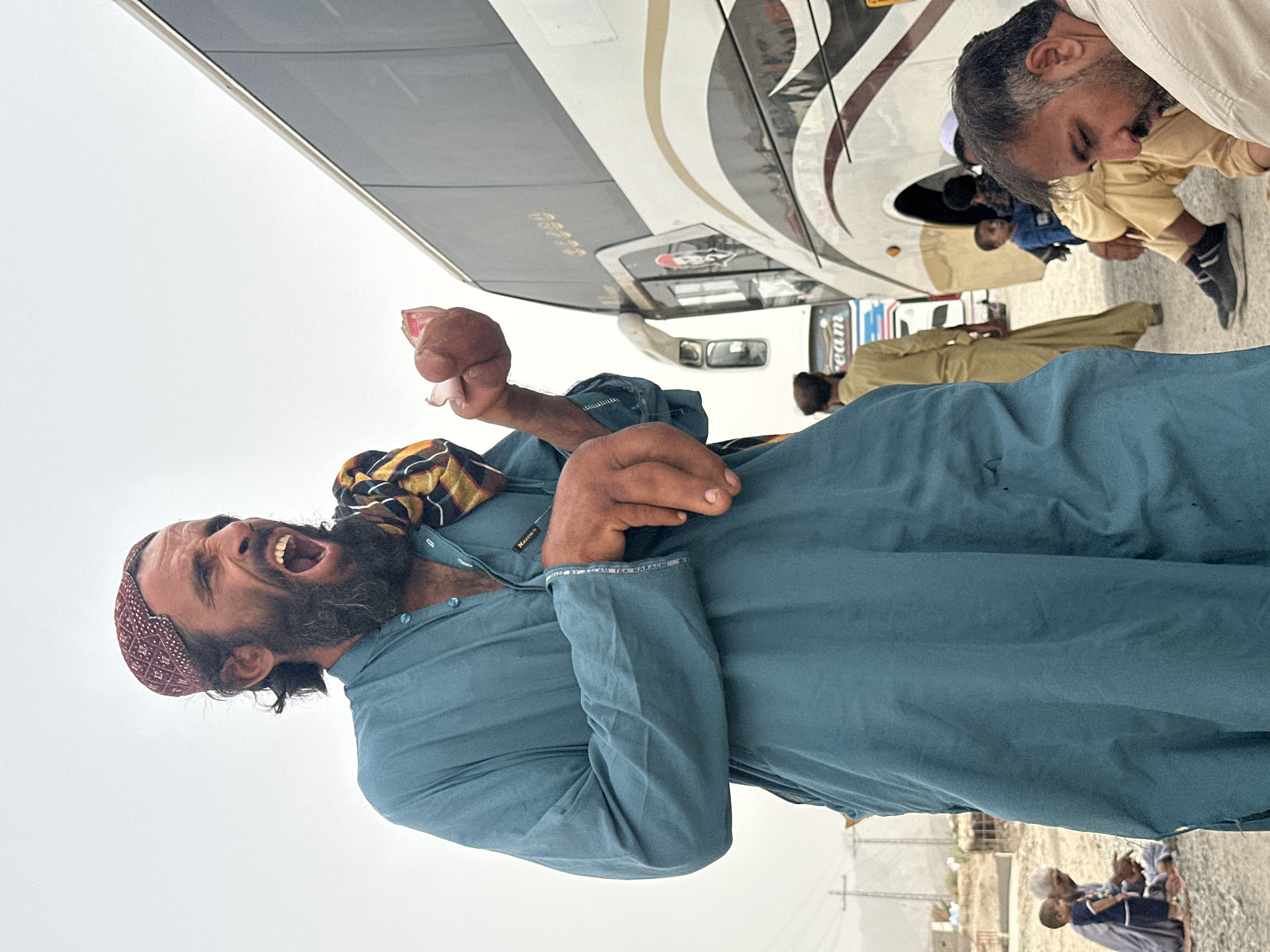 The media file depicts a group of people gathered outdoors, possibly near a bus. A man in the foreground is the primary subject, his head tilted back and mouth open in what appears to be a shout or laugh. He has a dark beard and is wearing a patterned cap and a teal shalwar kameez. His left hand, holding a small pink object, is raised near his face. Behind him, a bus with a distinctive white, grey, and brown design is visible. Other individuals are seen in the background, some seated near the bus. The setting appears to be a dry, possibly dusty, area with some low-lying structures and utility poles visible in the distance. The sky is overcast, suggesting daytime. The overall mood is energetic, with the foreground subject displaying a strong emotion. A partial view of text on the bus reads "team." Additionally, a label on the teal shalwar kameez of the foreground subject says "Nazmi 1959" and "MADE BY ADAM TEX KARACHI."