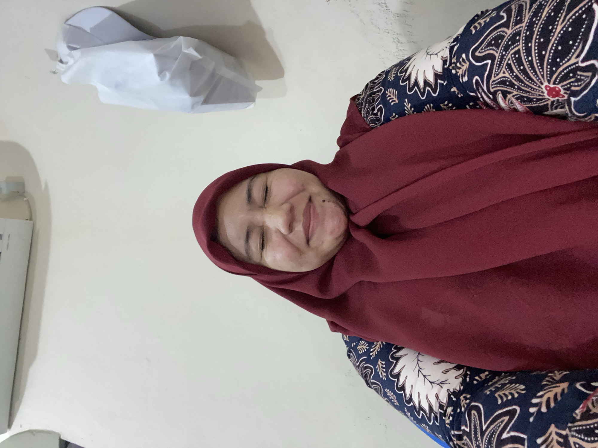 A medium shot shows a woman wearing a maroon hijab and a batik-patterned garment. Her eyes are closed, and she has a slight smile on her face. Her cheeks are plump, and there are some light freckles or skin blemishes visible on her face. The background is a plain, light-colored wall. Above and to the right of her head, a white object is hanging on the wall, possibly a piece of clothing or a bag. To the left of the woman, part of a white air conditioning unit is visible. The image appears to have been taken indoors, possibly in a residential setting in Banjarbaru, Indonesia.