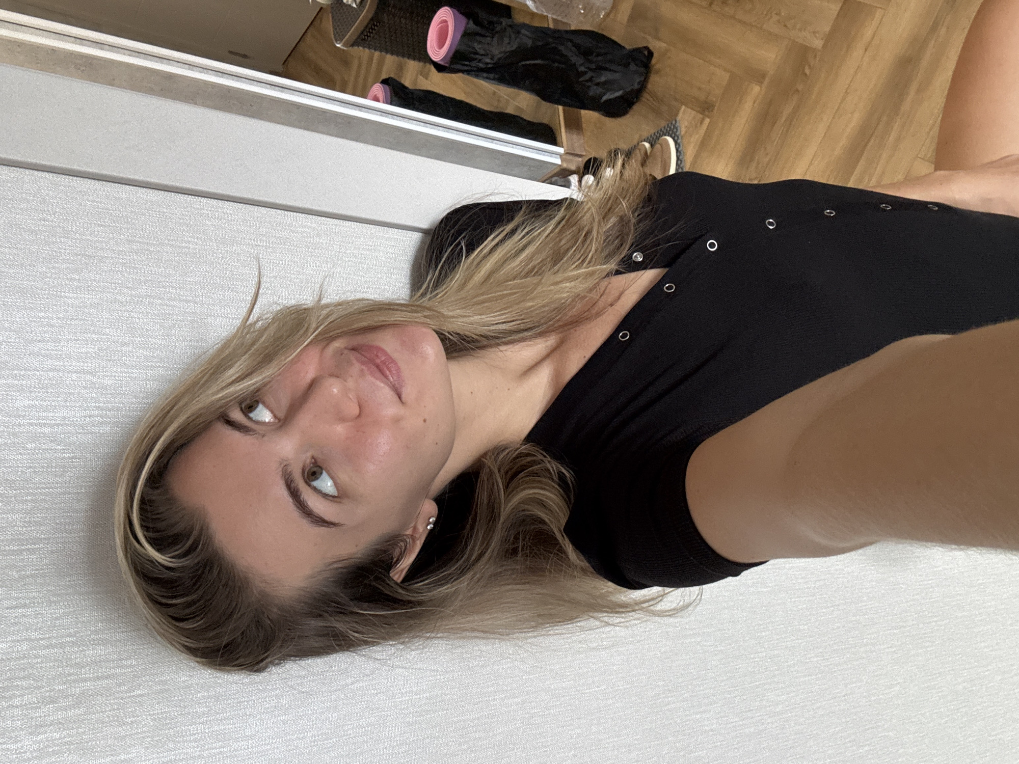 An unknown woman with blonde hair, wearing a black button-up shirt, poses for a selfie in an indoor setting. She is standing next to a wall and a doorframe, with wooden flooring visible. In the background, two rolled-up yoga mats in black bags are propped against a mirror. The woman is looking up and to the side, with a slight smile. Her left arm is extended as if holding the phone for the selfie.