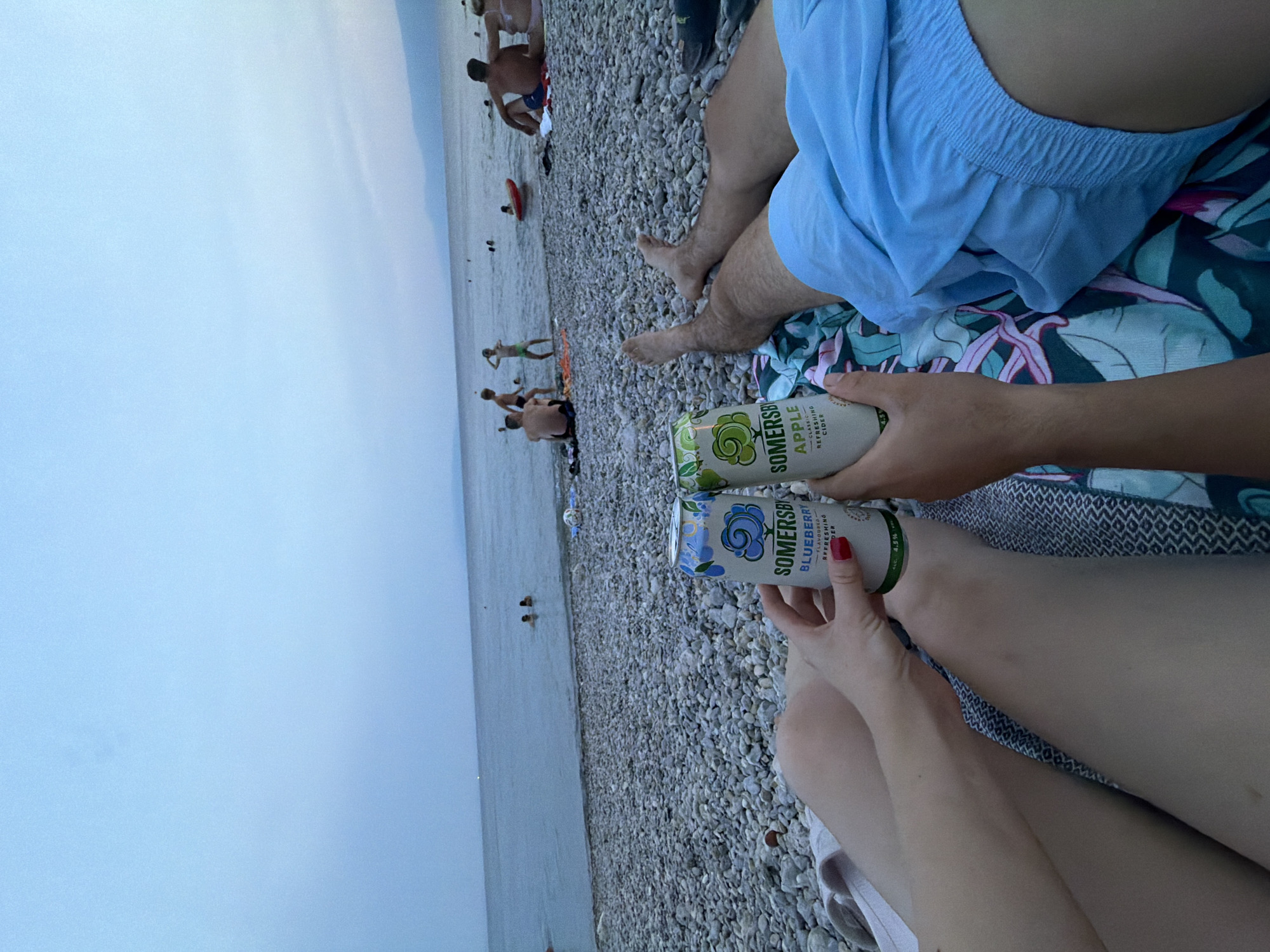 Two people are holding cans of Somersby cider on a pebble beach with the sea in the background. The sky is overcast and a few people are visible in the water and on the shore in the distance. One can is Somersby Blueberry and the other is Somersby Apple. The person holding the blueberry can has red nail polish. The person holding the apple can is wearing blue shorts.