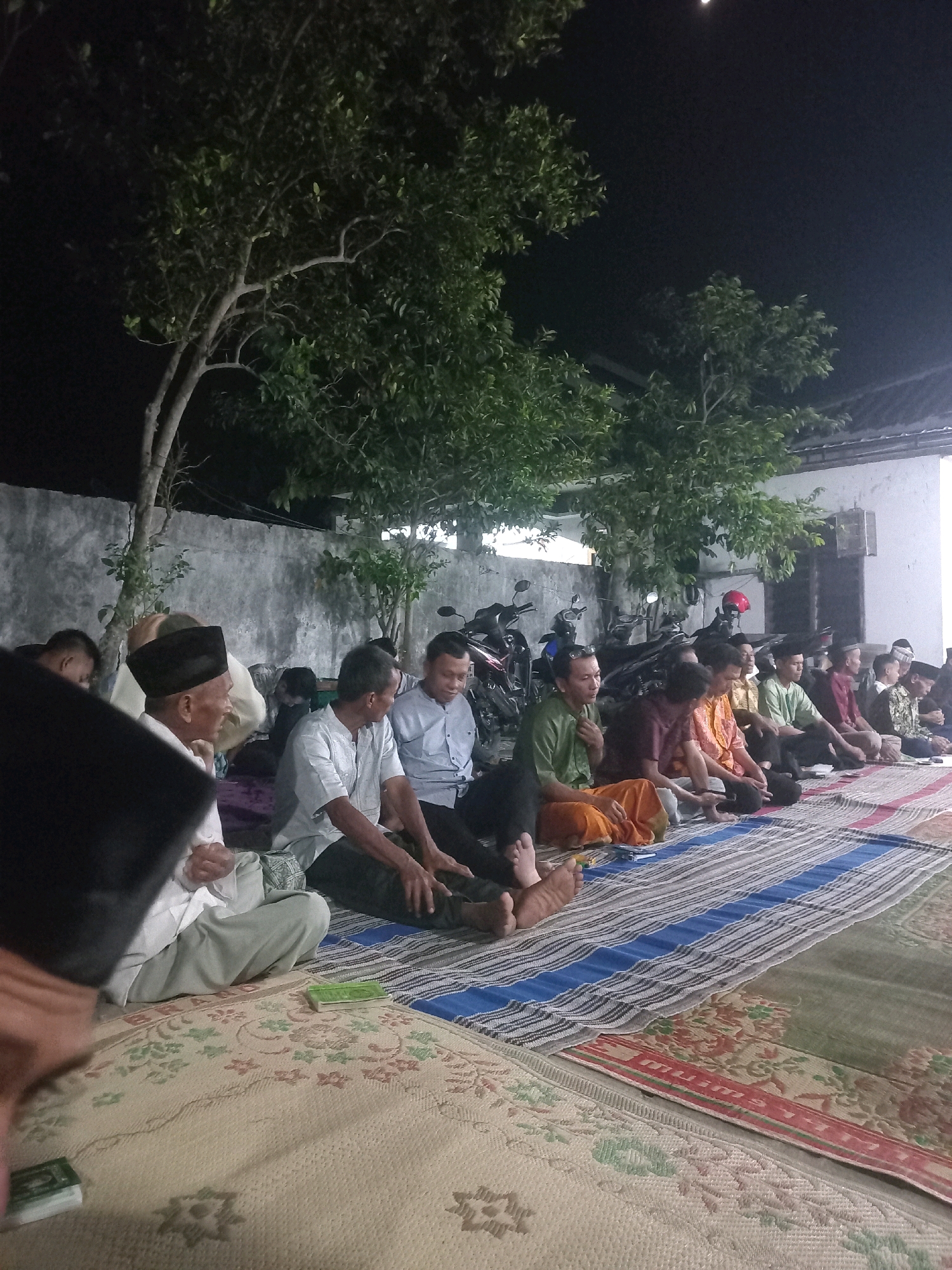 In Gambiranom, Indonesia, at night, a group of men are seated on mats in an outdoor gathering, likely a religious or community event. The scene is illuminated by artificial light, casting shadows and highlighting the dimly lit surroundings. The men are dressed in traditional attire, including peci caps and sarongs. They are seated in rows on a patterned mat. In the background, motorcycles are parked, and trees with foliage are visible against a dark sky. The environment suggests a modest residential or communal space.