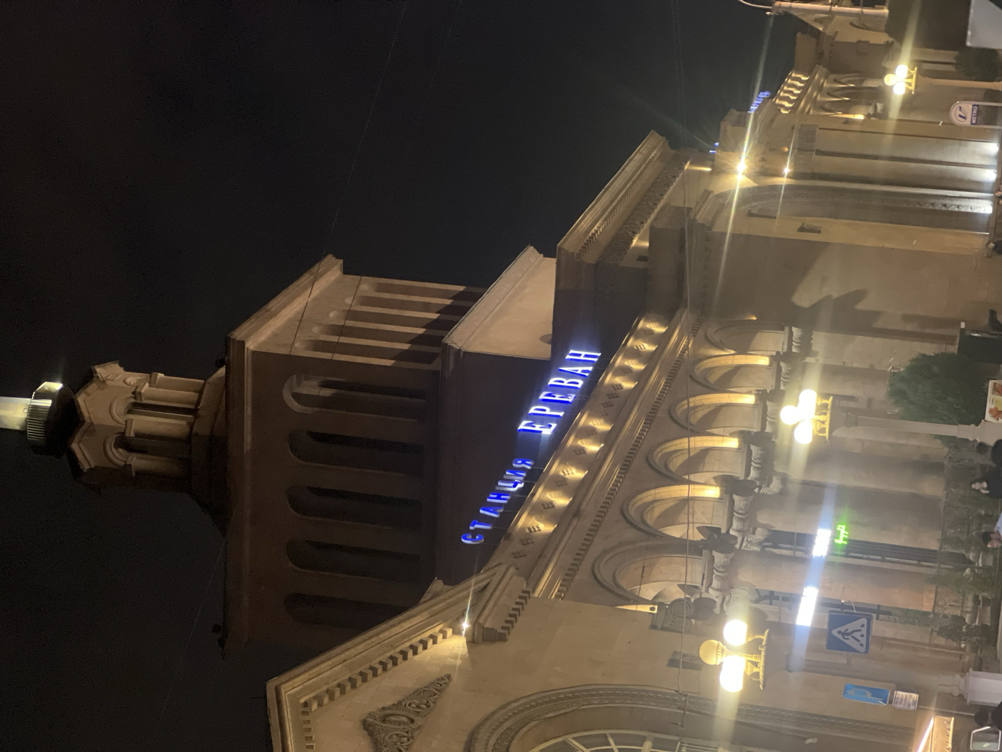 The image shows the Yerevan Railway Station in Yerevan, Armenia, at night. The building is illuminated, and the sign "Станция Ереван" (Station Yerevan) is visible in blue neon letters. The architectural style appears to be Soviet-era monumental. Streetlights and a few other signs are visible in the foreground. There are a few people standing near the front of the building and some trees in pots.