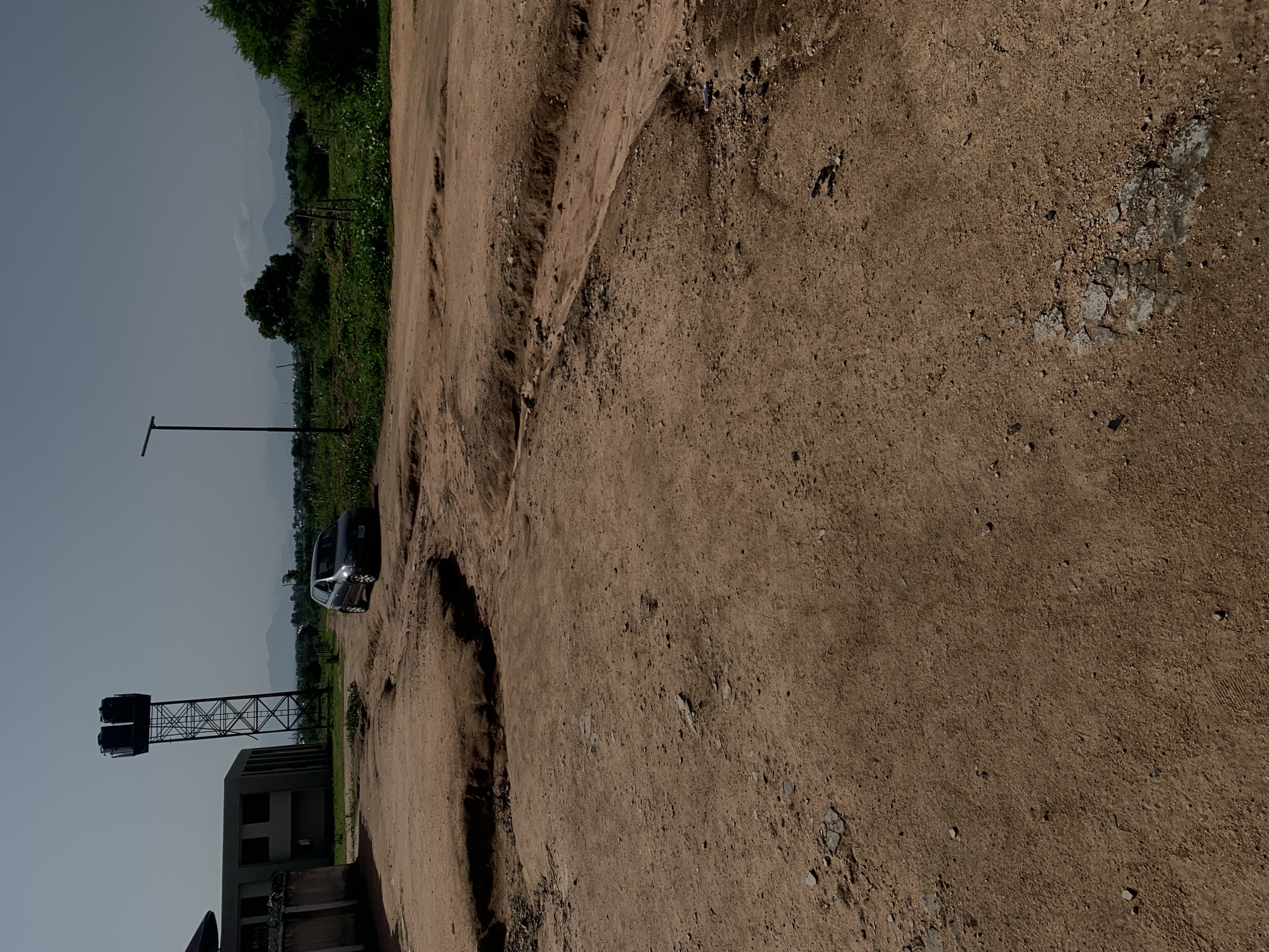 The image depicts an outdoor scene in Jalingo, Nigeria, characterized by a rough, unpaved dirt road with significant erosion and ruts, suggesting recent or ongoing water damage. A dark-colored car is parked on this road, facing away from the camera. To the left, a modern building with large windows is visible, along with a tall metal water tower topped with two tanks. In the background, a landscape of green vegetation and distant hills under a hazy sky is present. A solitary lamppost stands in the middle ground on the right side of the road. The overall environment appears somewhat arid, with the dirt road being a prominent feature.