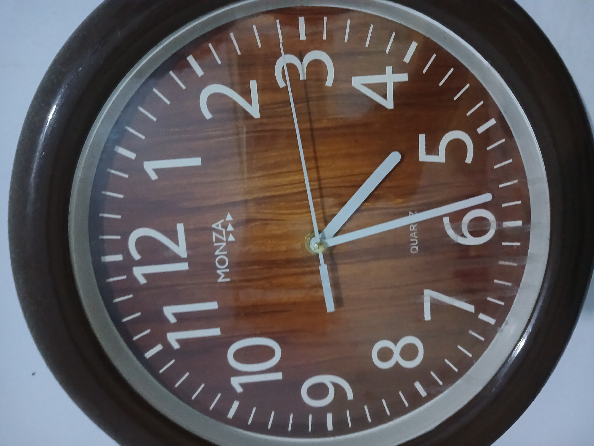 The image shows a close-up of a round analog wall clock with a wooden-patterned face. The clock has white Arabic numerals for hours and white markers for minutes. The hour hand is pointing between the 10 and 11, closer to the 11, and the minute hand is pointing towards the 4. The second hand is a thin white line pointing towards the 1. The clock is framed by a dark brown bezel. The background is a plain white wall. The location is a residence in Randuagung, Indonesia, likely a living room or dining area.
