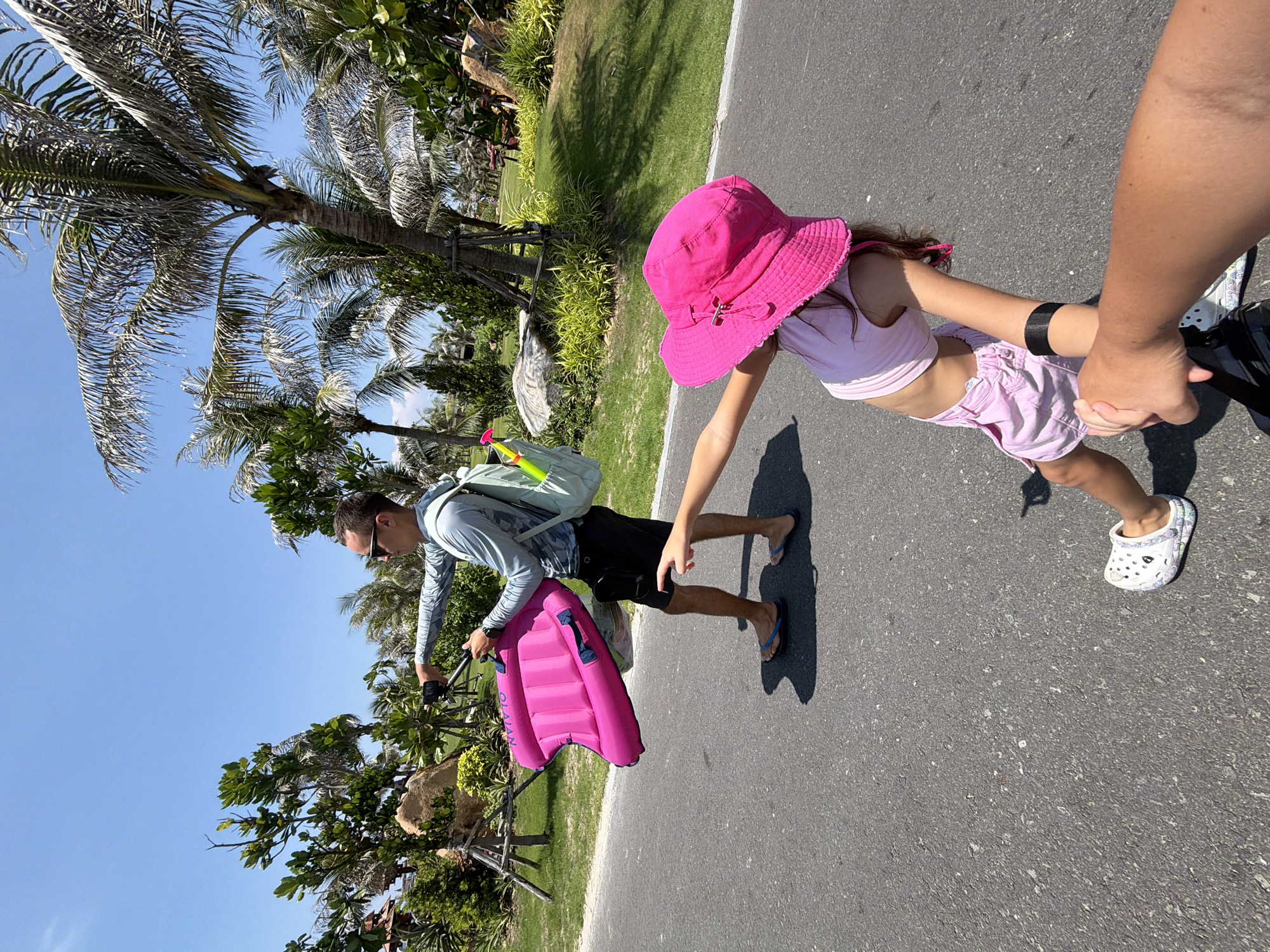 In Bac Cam Ranh Ward, Vietnam, a man with sunglasses and a light blue long-sleeved shirt walks on a paved path, carrying a pink inflatable float. He wears black shorts and blue flip-flops. A light blue backpack is slung over his shoulders, with a green water gun protruding from it. A young girl in a pink hat and pink shorts walks ahead of him, her arm extended as if holding his hand. The environment is tropical, with palm trees and lush greenery visible under a clear blue sky. The path appears to be part of a resort or park.