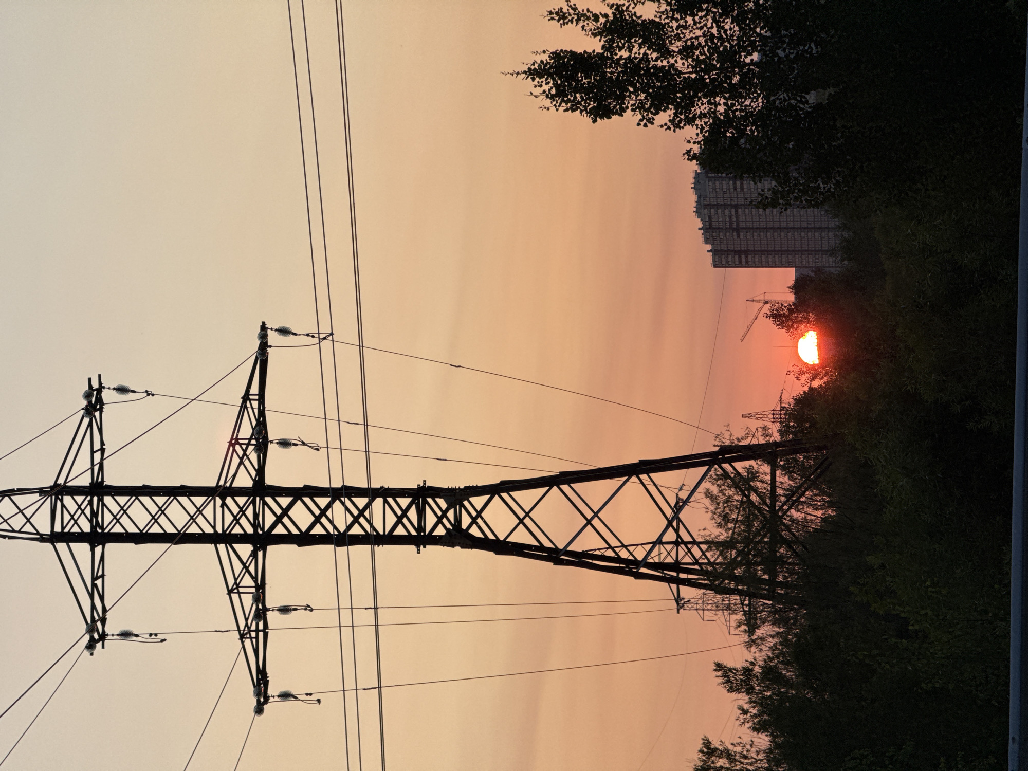 A sunset scene is captured with a large power line pylon prominently featured on the left side of the frame. The sky is a warm gradient of orange and pink hues, suggesting the sun is low on the horizon. In the background, a tall, multi-story building complex is visible, along with what appears to be a construction crane. Silhouetted trees and other vegetation occupy the middle ground, obscuring the lower parts of the pylons and the landscape. The overall impression is one of an urban or semi-urban environment at dusk. The specific location within the city of Surgut, Russia, is not visually identifiable from the image content alone. There are no people or discernible actions or interactions depicted.
