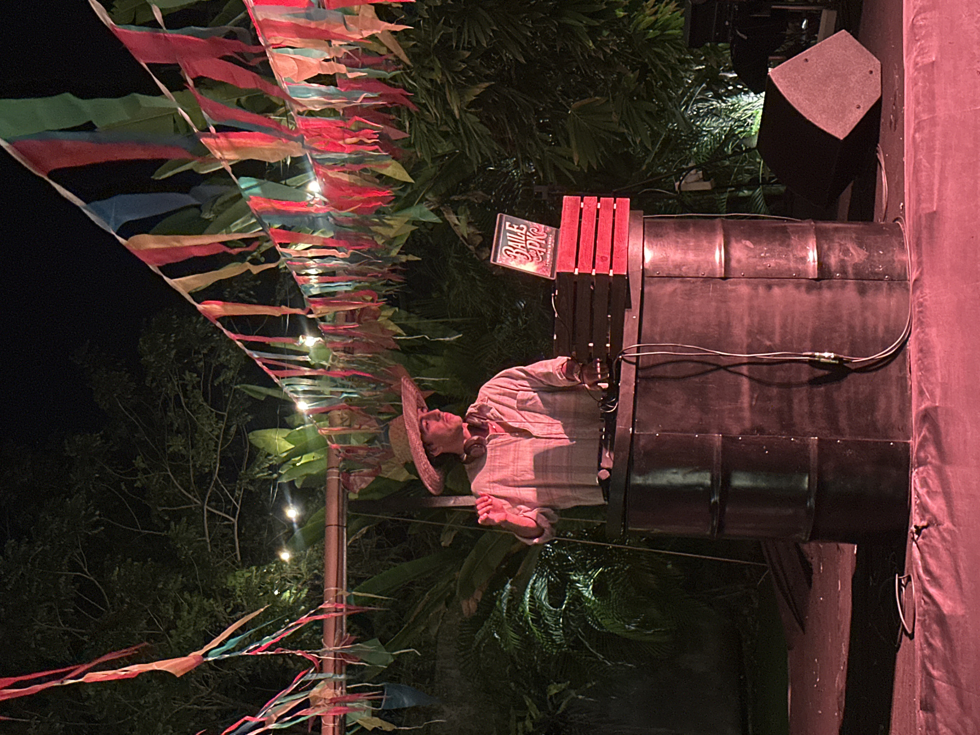 An outdoor event is taking place at night in Kuta Selatan, Indonesia, featuring a DJ. The DJ, a young man wearing a straw hat and a plaid shirt, is standing behind a DJ booth made from what appears to be a metal barrel. He is wearing headphones and is actively engaged with his equipment. In front of him, on the DJ booth, is a sign that reads "BAILE PKG" stacked on red wooden crates. Colorful flags are strung across the scene overhead, creating a festive atmosphere, with string lights also visible in the background. Lush green foliage surrounds the area, suggesting a tropical or garden setting. The ground in front of the DJ booth is covered in red fabric or lighting, casting a pink hue on the scene. A large speaker is positioned to the right of the DJ booth.