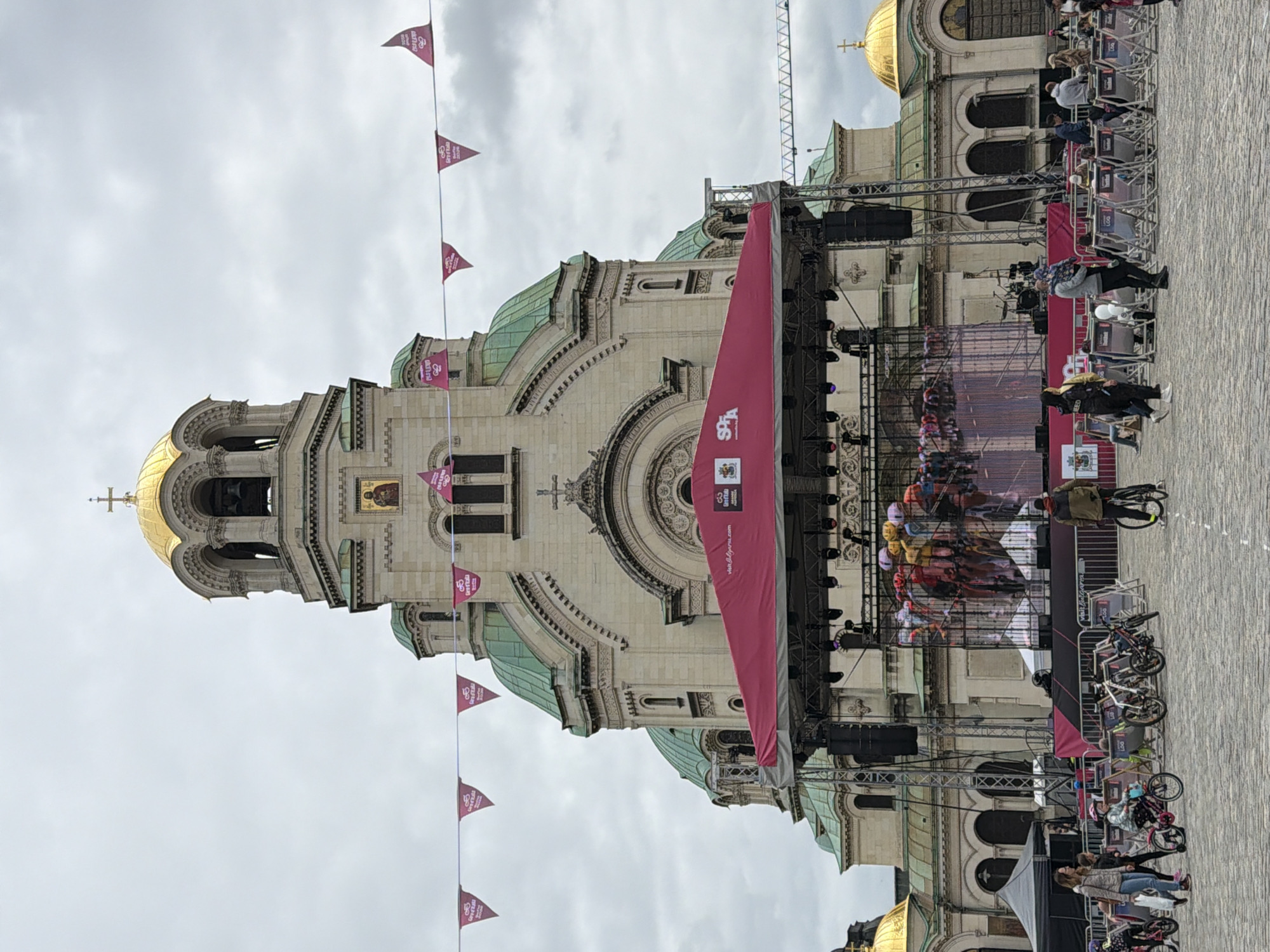 The image shows the Alexander Nevsky Cathedral in Sofia, Bulgaria. A stage has been set up in front of the cathedral, possibly for an event, with a large banner hanging on the side. Bicycles are parked near the stage. People are present, walking or standing in the square. The sky is overcast. There are pink flags hung on the poles.