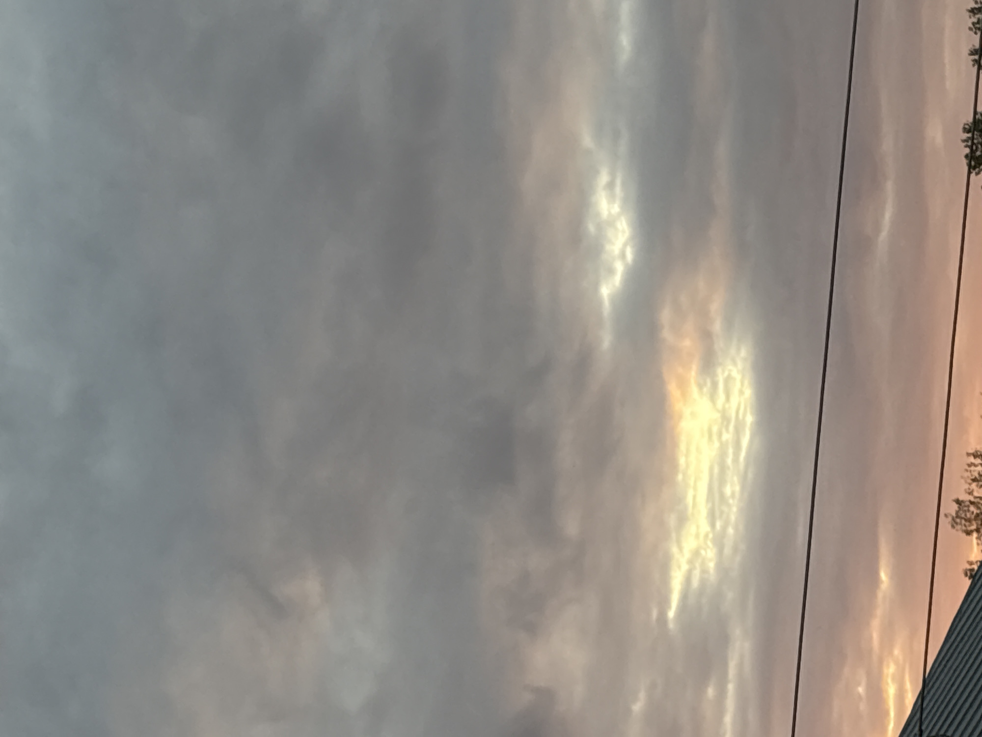 The image shows a cloudy sky with hints of sunset colors. A dark horizontal line, likely a power line, bisects the lower half of the image. Below the power line, a slanted dark roofline of a building is visible, along with silhouetted trees. The sky is predominantly gray, with some lighter, warmer tones of yellow and pink peeking through the clouds. The overall scene suggests an outdoor setting in a non-urban area, possibly on the outskirts of Georgiyevsk, Russia. There are no people or discernible actions depicted in the image.