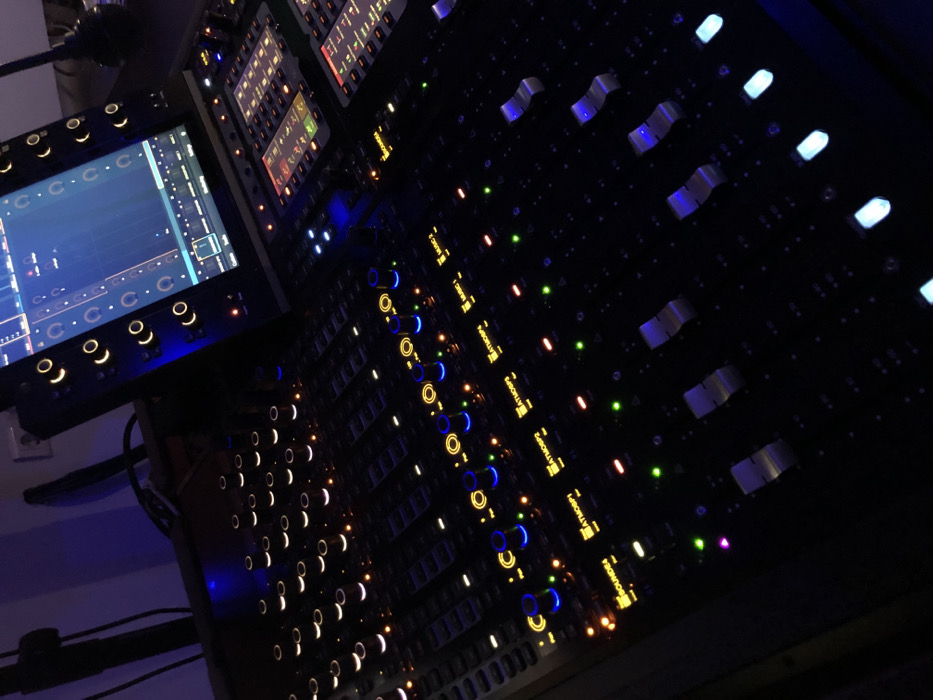 The image displays a brightly illuminated, dark-colored sound mixing console, likely situated in a professional audio studio or control room in Moschato, Greece. No people are visible, but the console is actively powered on, indicated by the glowing controls.
The console is adorned with numerous illuminated components, including blue-lit faders, yellow/orange-ringed knobs, and small green and red indicator lights. To the left, an integrated display screen shows a graphical user interface, featuring patterns of "C C C C" and "C C" across a blue background, possibly representing audio tracks, sequences, or control mappings.
The overall lighting is dim, highlighting the console's internal lights and suggesting either a low-light room setting or nighttime. The camera perspective is a dynamic, slightly elevated angle looking across the console. Visible text labels on the console's channels include "MAIN C 1", "ATMOSP1", "ATMOSP2", "ATMOSP3", "ATMOSP4", and "SOUNDS", indicating specific audio tracks or parameters. Other faint text and numerical readouts are visible on the various small displays and buttons. The scene conveys a professional and active audio production environment.