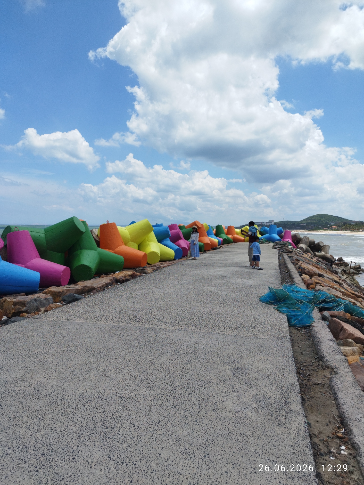 This media file depicts a scenic view of a breakwater adorned with vibrantly colored tetrapods. The tetrapods are arranged in a colorful gradient, featuring shades of blue, pink, orange, yellow, and green, creating a striking visual contrast against the natural surroundings. The breakwater itself is constructed from large rocks and concrete, with a paved walkway running along its length.
In the middle ground, a small group of people are visible on the walkway. A woman in a white top and long blue skirt stands near a cluster of tetrapods. Further down the walkway, a man with a child on his shoulders walks with another young boy beside him. The family appears to be enjoying a leisurely stroll along the coast.
The scene is set against a backdrop of a bright blue sky filled with fluffy white clouds, suggesting pleasant weather. The ocean is visible to the right, with gentle waves lapping against the shore. In the distance, a lush green hill forms a picturesque horizon. The overall atmosphere is one of tranquility and outdoor recreation.
A timestamp in the bottom right corner of the image reads "26.06.2026 12:29", indicating the date and time the photo was taken. This information, combined with the visual elements, suggests a daytime scene in Phu Thuy Ward, Vietnam, where the tetrapods serve as both functional coastal protection and an artistic display.