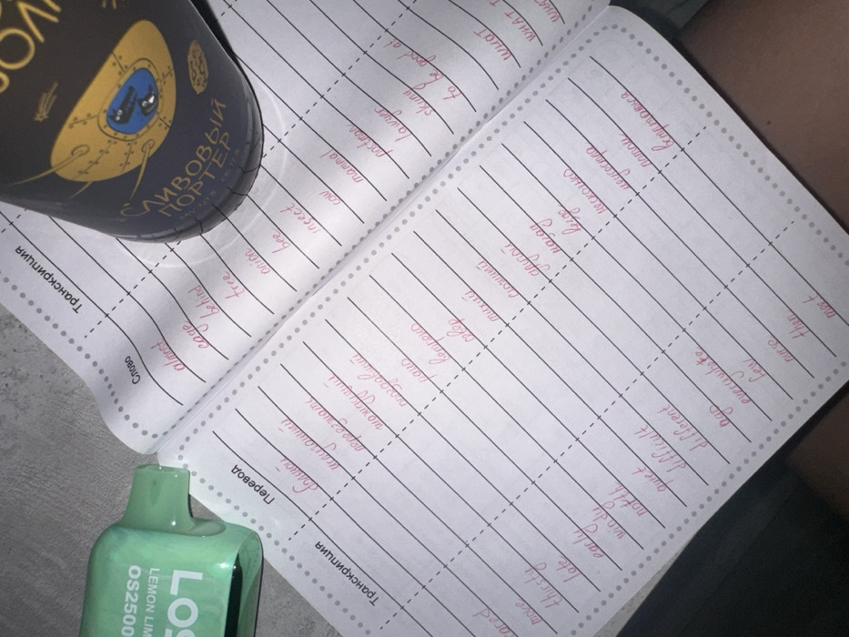 The image displays an open notebook with handwritten vocabulary words in Russian and English. On the left page, English words like "almost," "cage," and "bee" are translated into Russian. The right page shows words such as "большой," "испуганики," and "переезжать" being translated into English. A dark-colored bottle labeled "Сливовый Портер" and a green "LOS OS2500 LEMON LIME" disposable vape are visible on the notebook's left side.