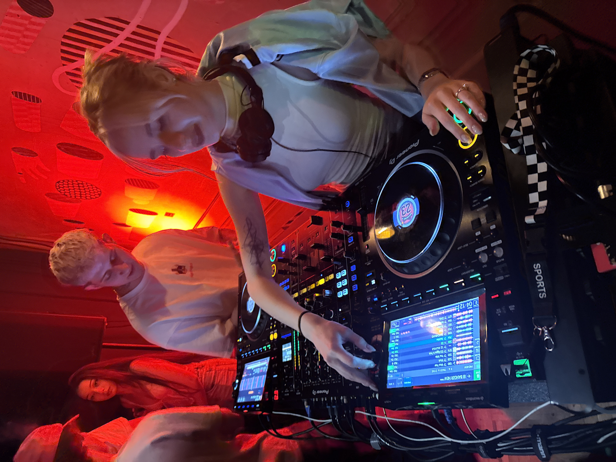 This dynamic photograph, taken in Sofia, Bulgaria, captures a high-energy electronic music event or club night under vibrant red lighting. The central figure is a young blonde woman, wearing a light-colored top and headphones around her neck, actively engaged in DJing. Her right hand, adorned with rings, is positioned on a Pioneer DJ controller, interacting with its illuminated buttons and screens. A tattoo is visible on her right forearm.
Behind and to the left of the DJ, a young blonde man in a white t-shirt observes the setup. A third person, a young woman with dark hair, is partially visible in the lower left background. The scene is illuminated predominantly by a strong red light, creating dramatic shadows and highlights, with abstract red patterns visible on the walls or ceiling. Green and blue lights emanate from the screens and buttons of the professional DJ equipment.
Visible text includes "Pioneer DJ" on the equipment, "SPORTS" on a black and white checkered strap, and various details on the screens such as "BEAT LOOP," "RELOOP/EXIT," "PREVIEW," "BANGER NEW***," "@rexorubox," "MV MINTO," and a time indicator "04:12," along with numerous track numbers and settings like "23," "704," "12," "700," "74," and "101." The image's tilted perspective adds to the energetic and immersive feel of the performance.