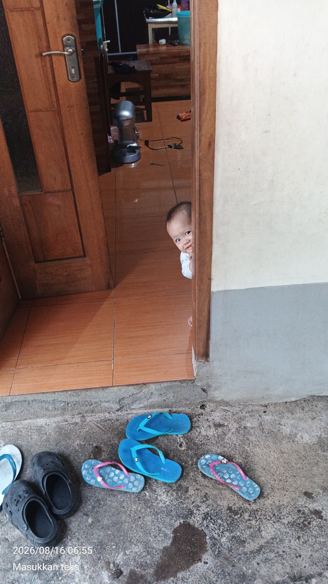 A baby peeks out from behind a wooden doorframe in what appears to be a residential setting. The baby, wearing a white shirt, is positioned on a tiled floor, with the door slightly ajar. In the background, a silver kitchen appliance is visible on the floor. Outside the door, on a concrete surface, there are several pairs of flip-flops and clogs scattered about. The scene suggests a casual, domestic environment.