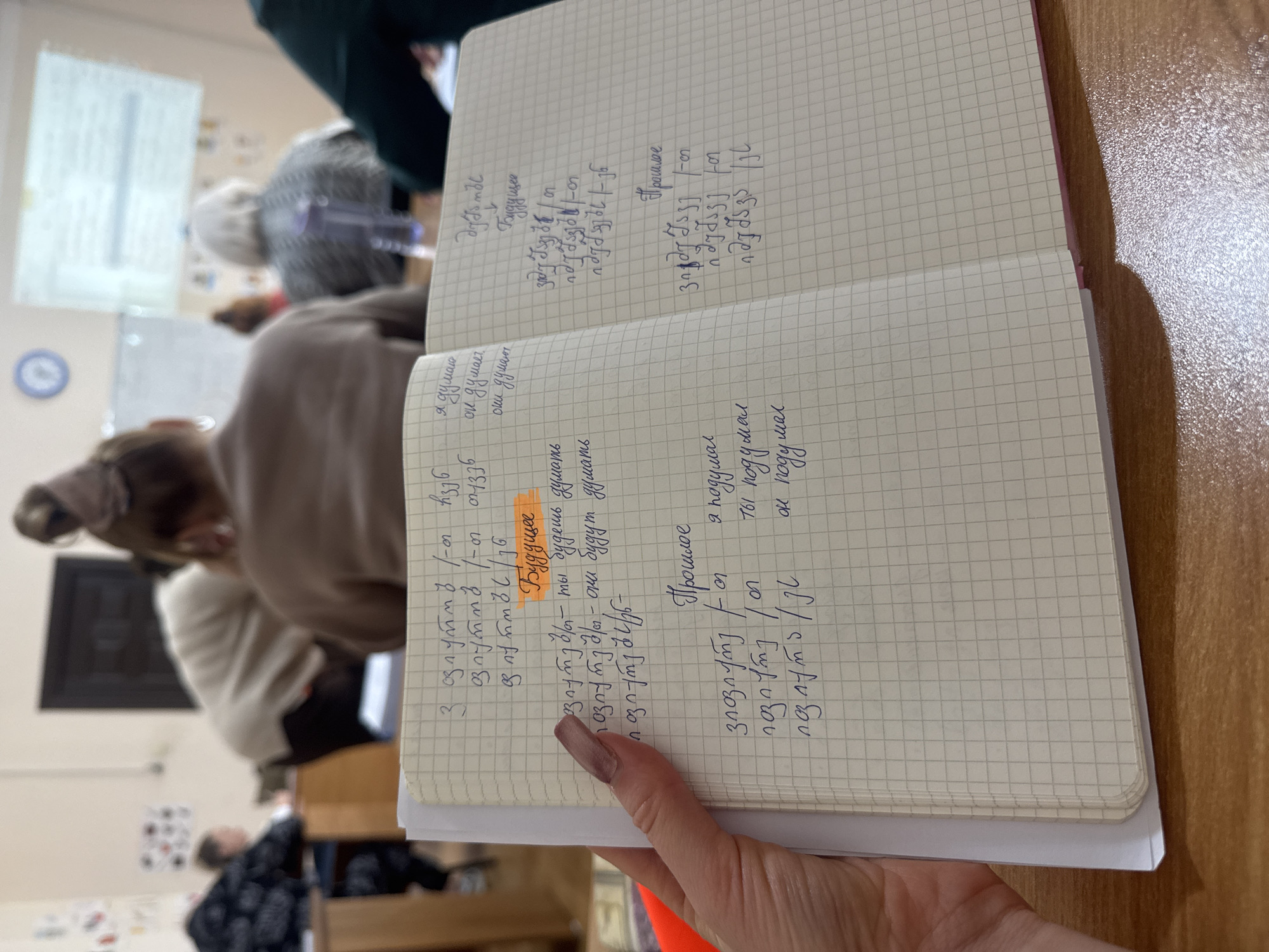 A first-person perspective shows a left hand with manicured, light-colored nails holding open a grid-lined notebook over a polished wooden desk. The notebook contains handwritten notes in Georgian script and Cyrillic (Russian), detailing verb conjugations for "think" in future ("Будущее") and past ("Прошлое") tenses, indicating a language lesson. The background, out of focus, reveals an indoor classroom or lecture hall in the city of Unknown, Unknown. Several individuals are seated at wooden desks, facing forward. Visible attendees include a person with light brown hair pulled back, and an older individual with white hair wearing a grey patterned top, both viewed from behind. A clear plastic water bottle rests on a desk in the mid-right background. A projector screen displaying blurred text and a blue circular wall clock are mounted on the far wall. Another person in a light-colored top is observed leaning over a desk in the mid-left background. The scene depicts an active educational setting from the viewpoint of a note-taker.
