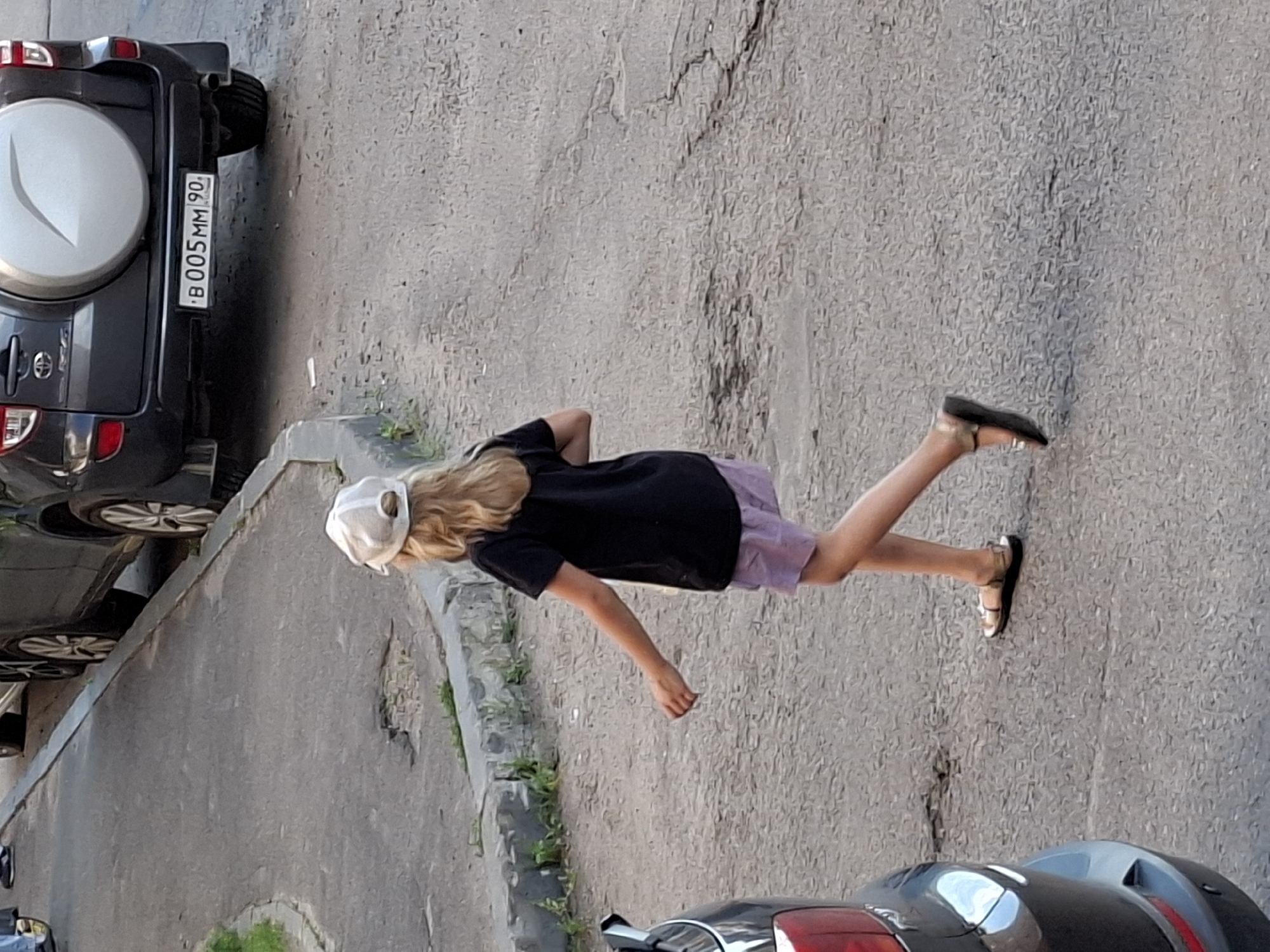 In an outdoor urban setting, a young person with blonde hair, wearing a white baseball cap, black t-shirt, and purple shorts, walks across a paved road. The person is seen from behind and to the right, with their arms slightly bent and hands open. The ground is asphalt with visible cracks and patches. To the right of the person, a dark-colored SUV is parked, displaying a license plate that reads "B 005 MM 90". To the left of the person, the rear of a dark-colored car is visible. The background shows a sidewalk with a stone border and some green vegetation. The lighting suggests it is daytime.