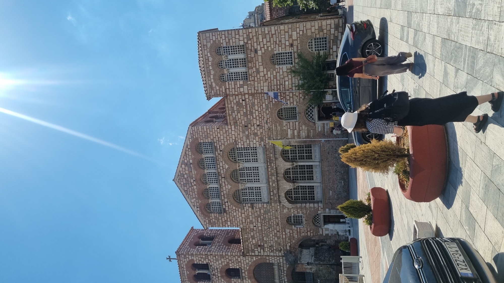 The image shows a view of the Church of Saint Demetrius in Thessaloniki, Greece. The sun is shining brightly, casting long shadows on the paved ground. In the foreground, two women are walking away from the camera. One woman wears a white hat, a black and white checkered shirt, and a black skirt. The other woman wears a burgundy top and grey pants. A dark grey car is parked to the right, and a black car is partially visible on the left. Several red planters with small trees are placed around the plaza. The church facade, constructed of stone and brick, features numerous arched windows and multiple levels. A cross is visible atop one of the bell towers.