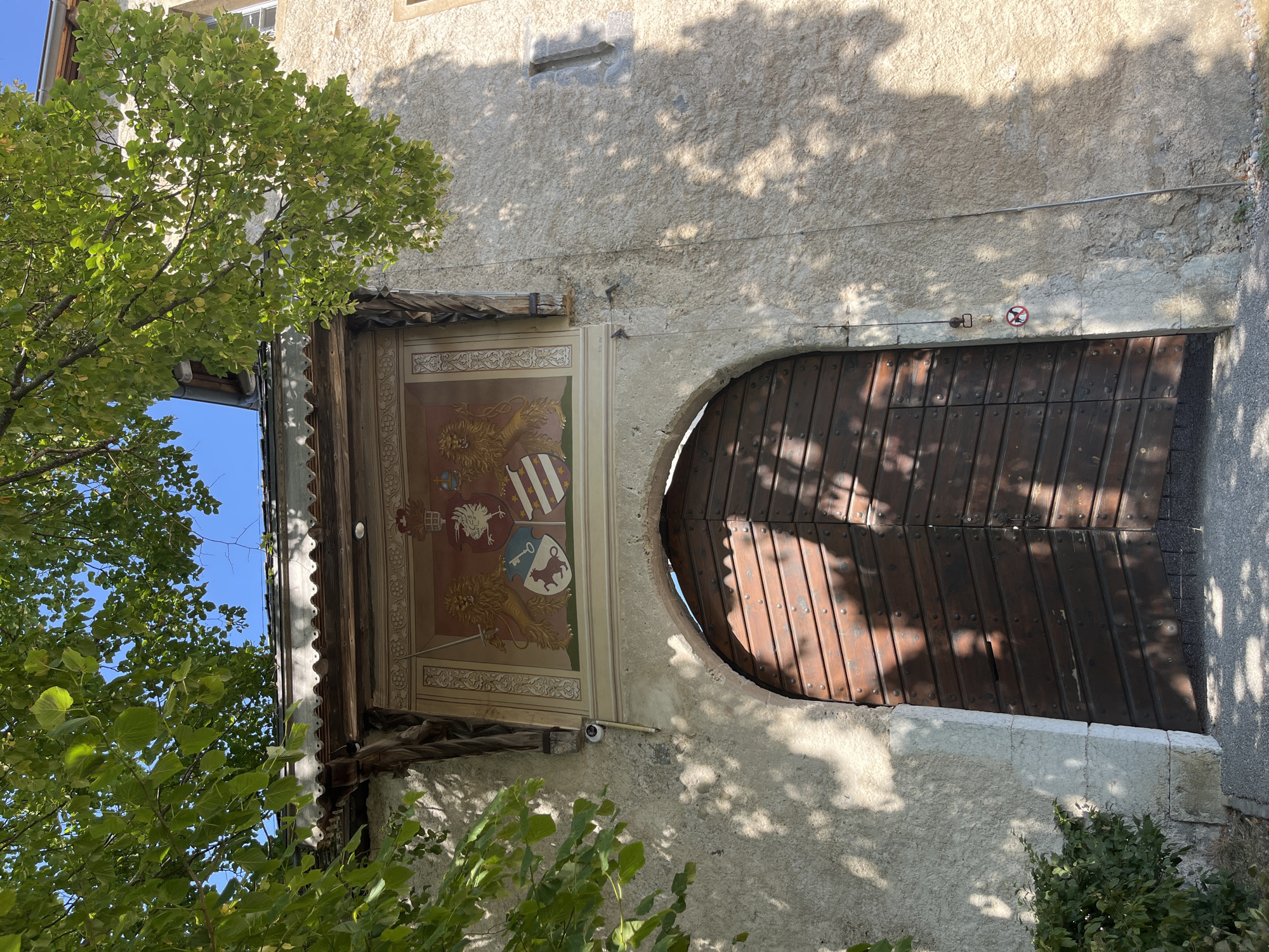 The image shows a large wooden double door set within a stone archway. Above the archway is a mural depicting a coat of arms with two lions and a crown. The building appears to be made of stone and plaster with a tiled roof. Trees with green leaves overhang the scene, casting shadows. A security camera is mounted on the wall to the left of the mural. To the right of the door, a circular sign with a red border shows a silhouetted animal with a red diagonal line through it, indicating no pets. The scene is outdoors, under a bright sky. The location of Epagny, Switzerland is not visible in the image.