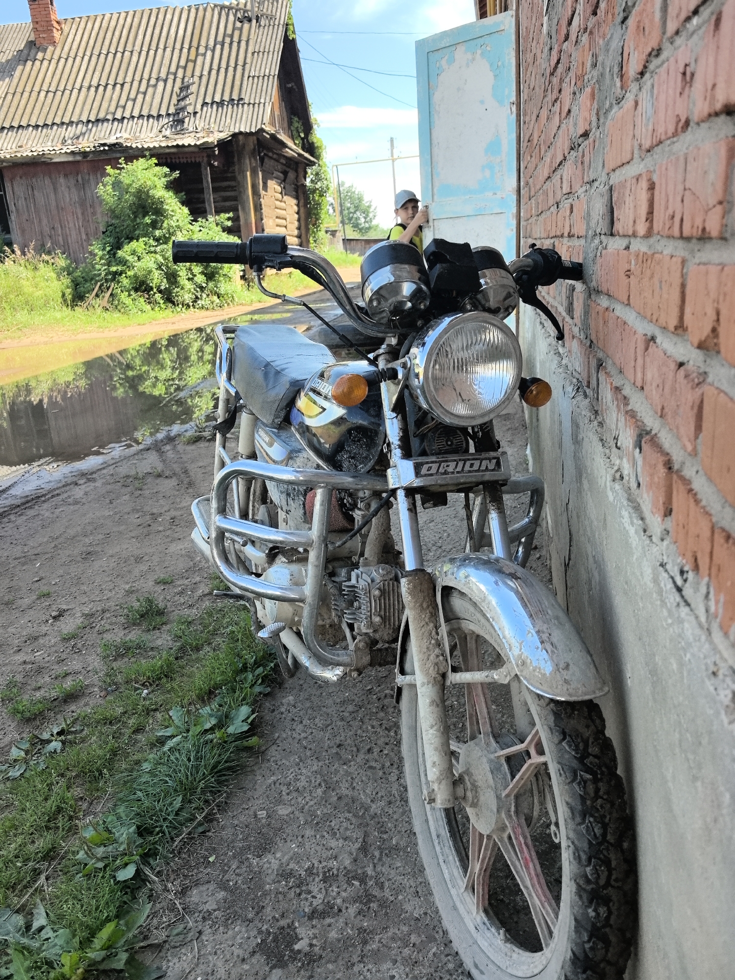 A motorcycle is parked next to a brick wall in Noviy, Russia. The motorcycle is dirty and has a metal frame. A person wearing a yellow shirt and a grey cap is standing in the background near a blue door. A wooden house with a grey roof is visible behind the motorcycle. There is a puddle of water on the ground in front of the house. The sky is blue with some clouds.
