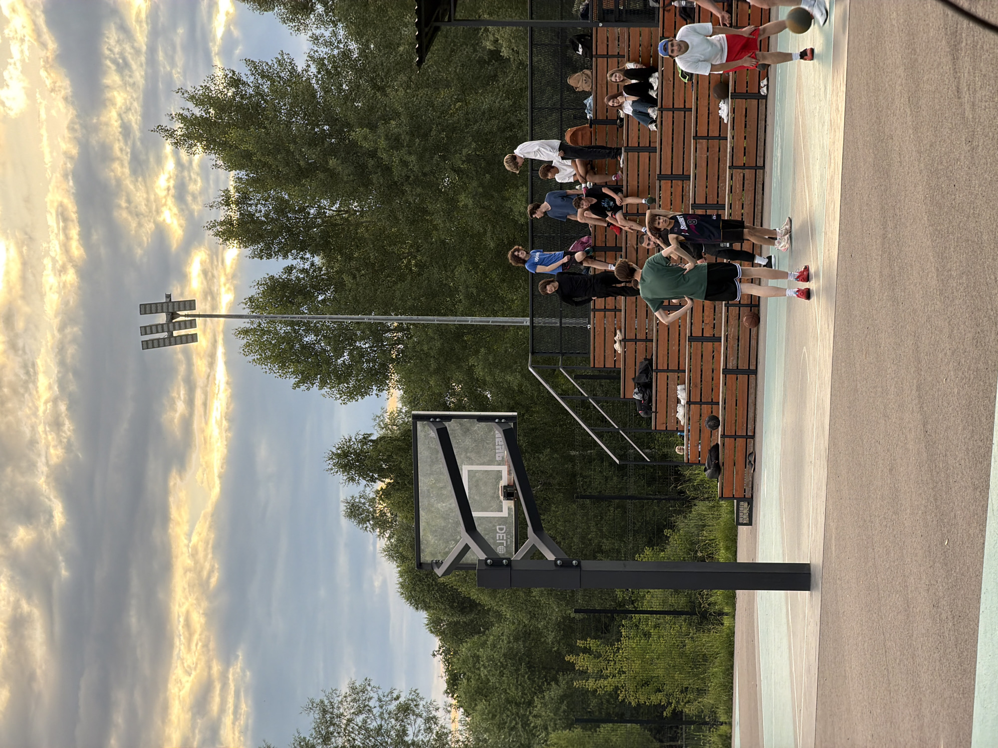 The image captures a group of young people gathered on and around a basketball court. The scene appears to be outdoors, likely in a park or recreational area, with lush green trees surrounding the court and in the background. A prominent basketball hoop with a glass backboard is visible on the left side of the frame. Wooden bleachers, occupied by several individuals, are situated behind the court, suggesting a viewing area or a place to rest.
The people on the court and bleachers are dressed casually, in athletic wear and everyday clothing. Some are standing, while others are seated. One individual on the court is actively engaged with a basketball, suggesting a game or practice is underway. The overall mood seems relaxed and social, with a sense of camaraderie among the group.
The lighting indicates it is either early morning or late afternoon, with the sun casting a warm glow. The sky is partially cloudy, with streaks of yellow and white clouds against a pale blue backdrop, creating a dynamic sky.
There is visible text on the basketball backboard, reading "DEFECT". The location context is provided as Kazan, Russia, though no specific landmarks within Kazan are identifiable in the image. The scene suggests a casual basketball gathering or a break during a game.