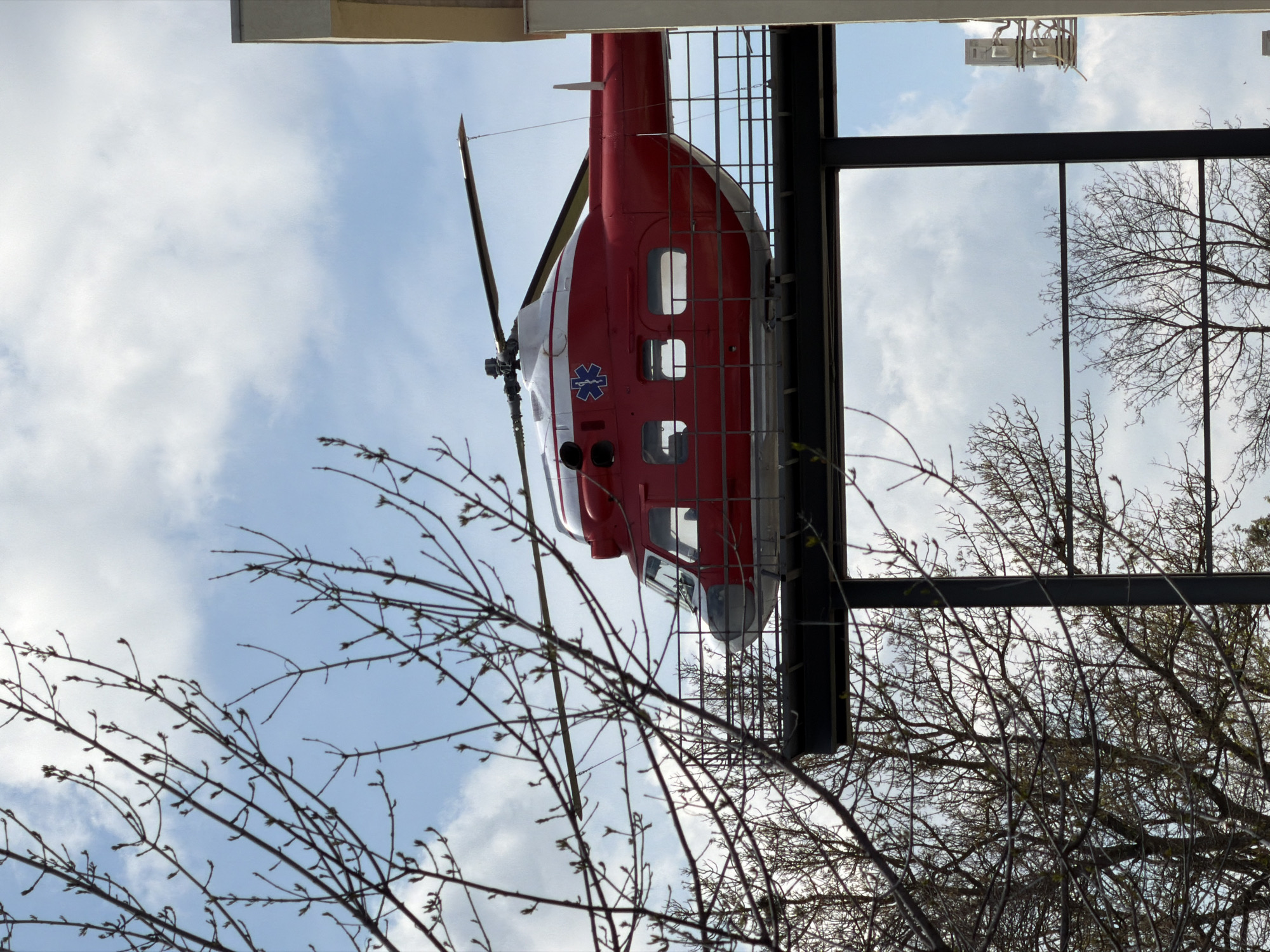 A red and white helicopter, possibly a medical transport, is mounted on a black metal frame outside a building. The helicopter is partially obscured by bare tree branches in the foreground. The background shows a cloudy sky and parts of other trees. The location is in Pleven, Bulgaria.