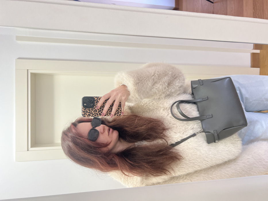 The image presents a mirror selfie of a female individual taken indoors within a residence in Budva, Montenegro. The subject, possessing long, reddish-brown hair, wears a light beige faux fur or fluffy jacket, light blue trousers, and dark, rectangular sunglasses. Her right hand holds a smartphone with a leopard print case, directed at the mirror to capture her reflection. A dark grey, structured handbag with twin top handles and a shoulder strap is worn diagonally across her body, resting on her left hip. The background shows an interior space with white walls and a light-colored door frame. A partial view of a wooden floor is visible in the reflection on the right side of the image.