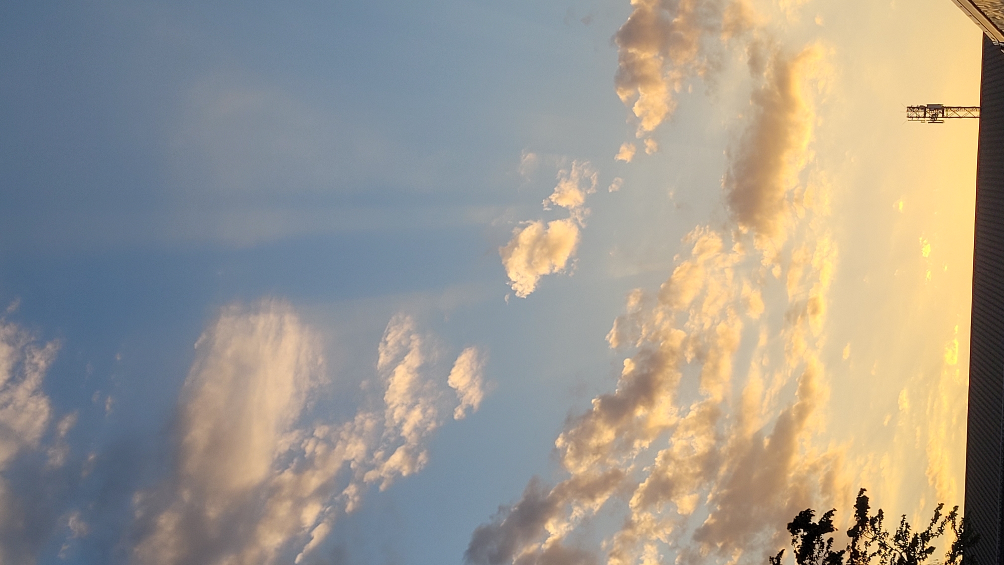 This media file depicts a dramatic sunset sky over Hengersberg, Germany. The scene is dominated by a vast expanse of sky, with fluffy, golden-tinged clouds scattered across a fading blue canvas. The clouds are illuminated by the setting sun, casting warm hues of orange and yellow, contrasting with the cooler blue of the upper atmosphere. Faint sunbeams are visible, adding depth and texture to the sky.
On the right side of the frame, a silhouette of a building with a textured roofline suggests an urban or industrial setting. Rising above this, a section of a construction crane is visible against the bright sky, hinting at ongoing development or infrastructure work in the area. At the very bottom right, the dark silhouette of tree branches adds a natural element to the scene, framing the lower edge of the composition.
The time of day is clearly late afternoon or early evening, given the intense golden light and the sunset colors. The weather appears clear, with the clouds indicating typical atmospheric conditions for a sunset. There are no people visible in the image, and no text is present. The overall mood conveyed is one of natural beauty and transition, as day gives way to night.