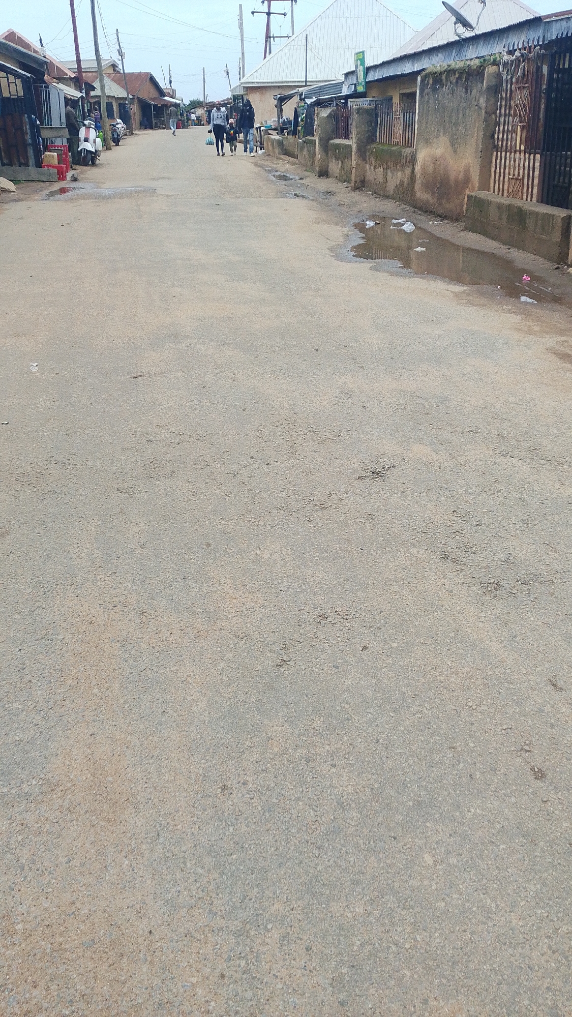 A low-angle, eye-level, full shot captured on an overcast day shows a paved road lined with buildings on both sides. The road appears to be in a residential or commercial area with modest structures. On the left side of the road, there are shops with open fronts and a white scooter parked. Further down the road, a few people are walking. On the right side, the buildings have a more traditional appearance with mud walls and corrugated iron roofs, some with metal gates. Puddles of water are visible on the road and near the buildings, suggesting recent rain. The sky is uniformly gray, and there are utility poles and wires visible in the background. The scene suggests an everyday street in a developing area.