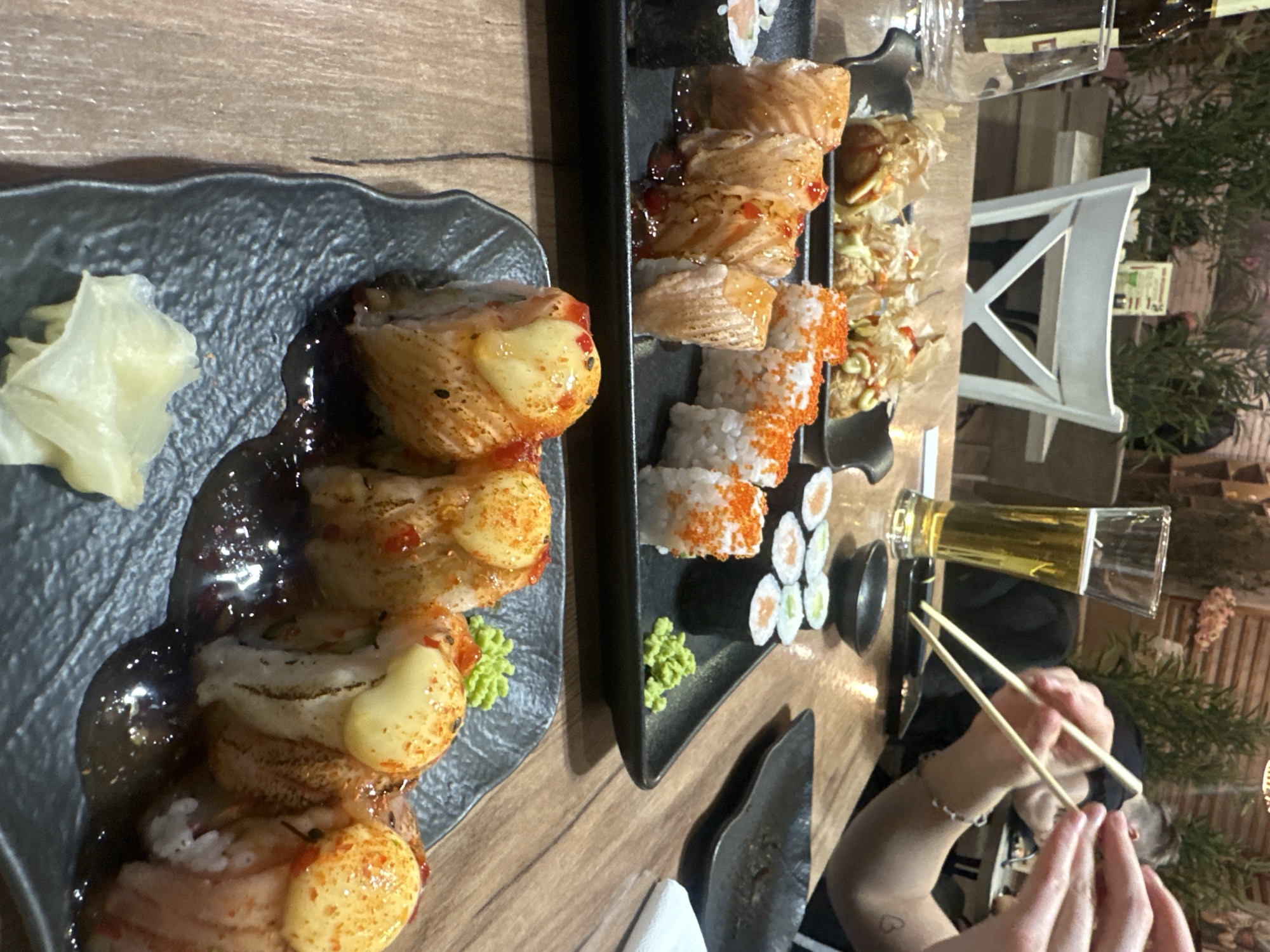 A table is set with two plates of sushi, a beer glass, and chopsticks. One plate has a stack of sushi rolls with a dark sauce, and a side of pickled ginger. The other plate contains a variety of sushi rolls, including salmon rolls, California rolls, and small seaweed rolls, with a dollop of wasabi. A person is using chopsticks, with their arm visible. The setting suggests an urban environment, possibly a restaurant in Varna, Bulgaria. There's a white chair and some greenery in the background. The image focuses on the food and the act of eating sushi.
